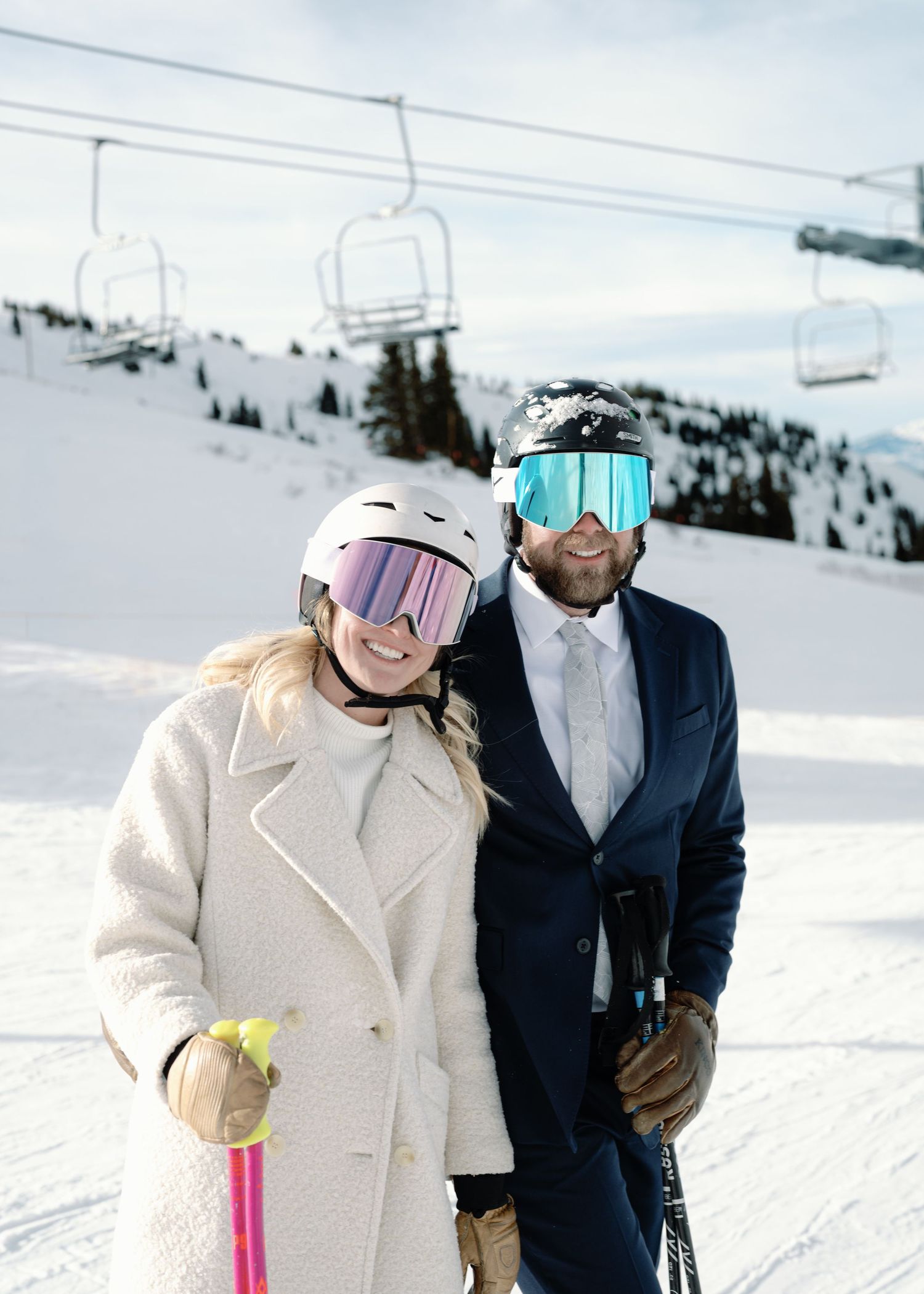 Bride and groom in ski helmets and goggles smile together beneath chairlift on snowy ski slope.