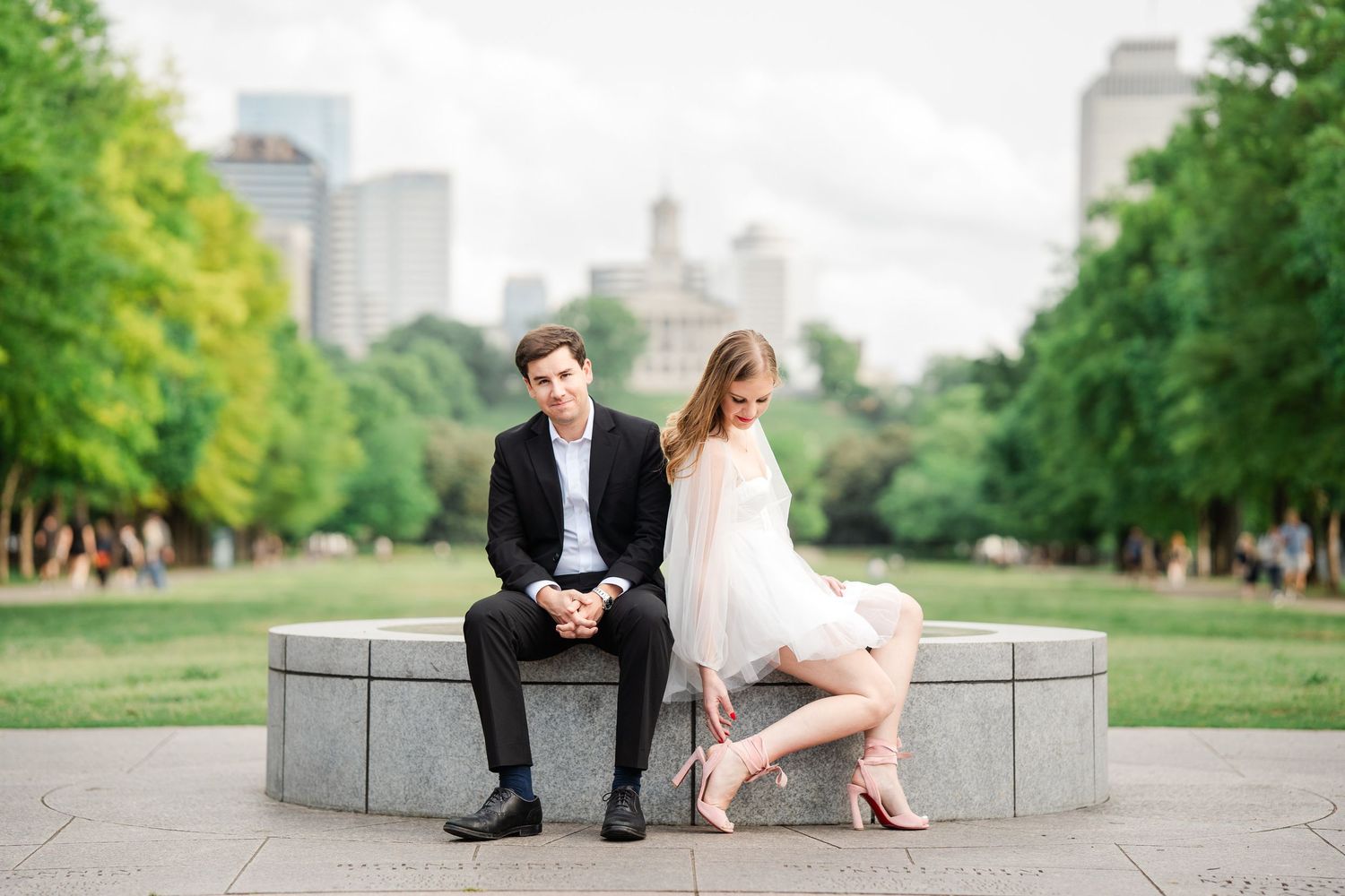 Bride and groom sitting back to back on a stone bench in a lush green park with city skyline.