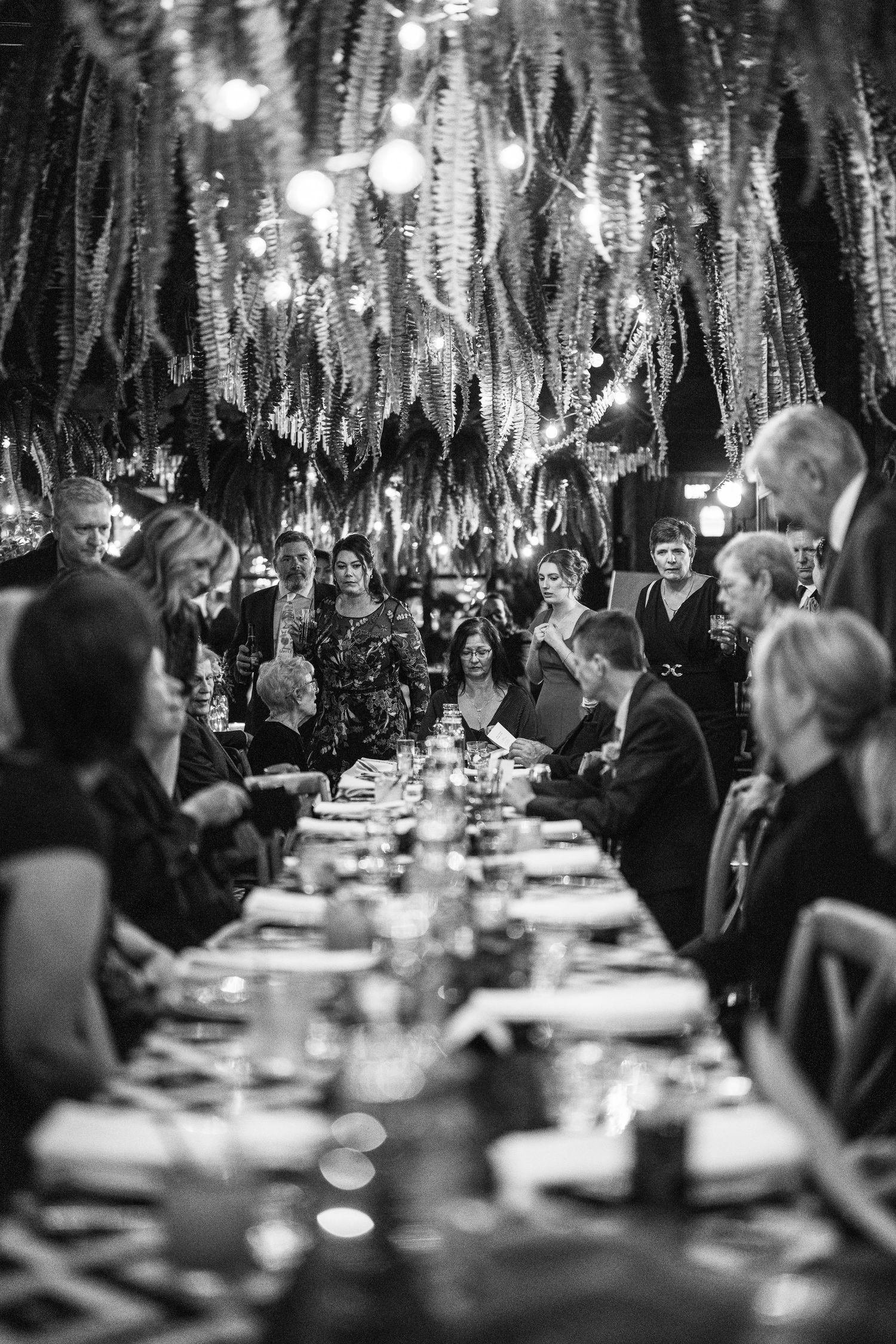 Black and white photo of wedding guests gathered around a long decorated table under hanging ferns and string lights.