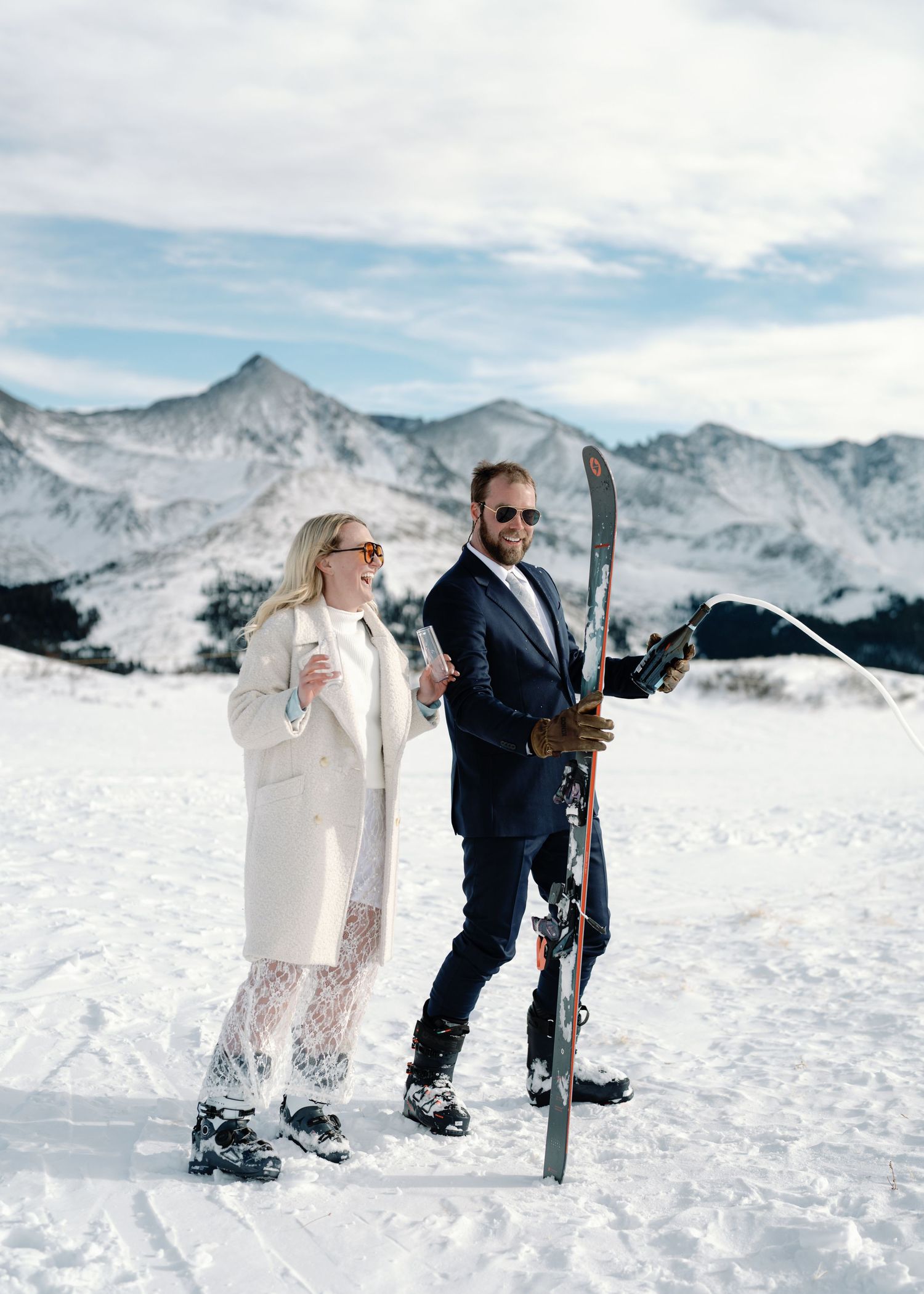 Couple in wedding attire holding skis on a snowy mountain slope with peaks in the background.