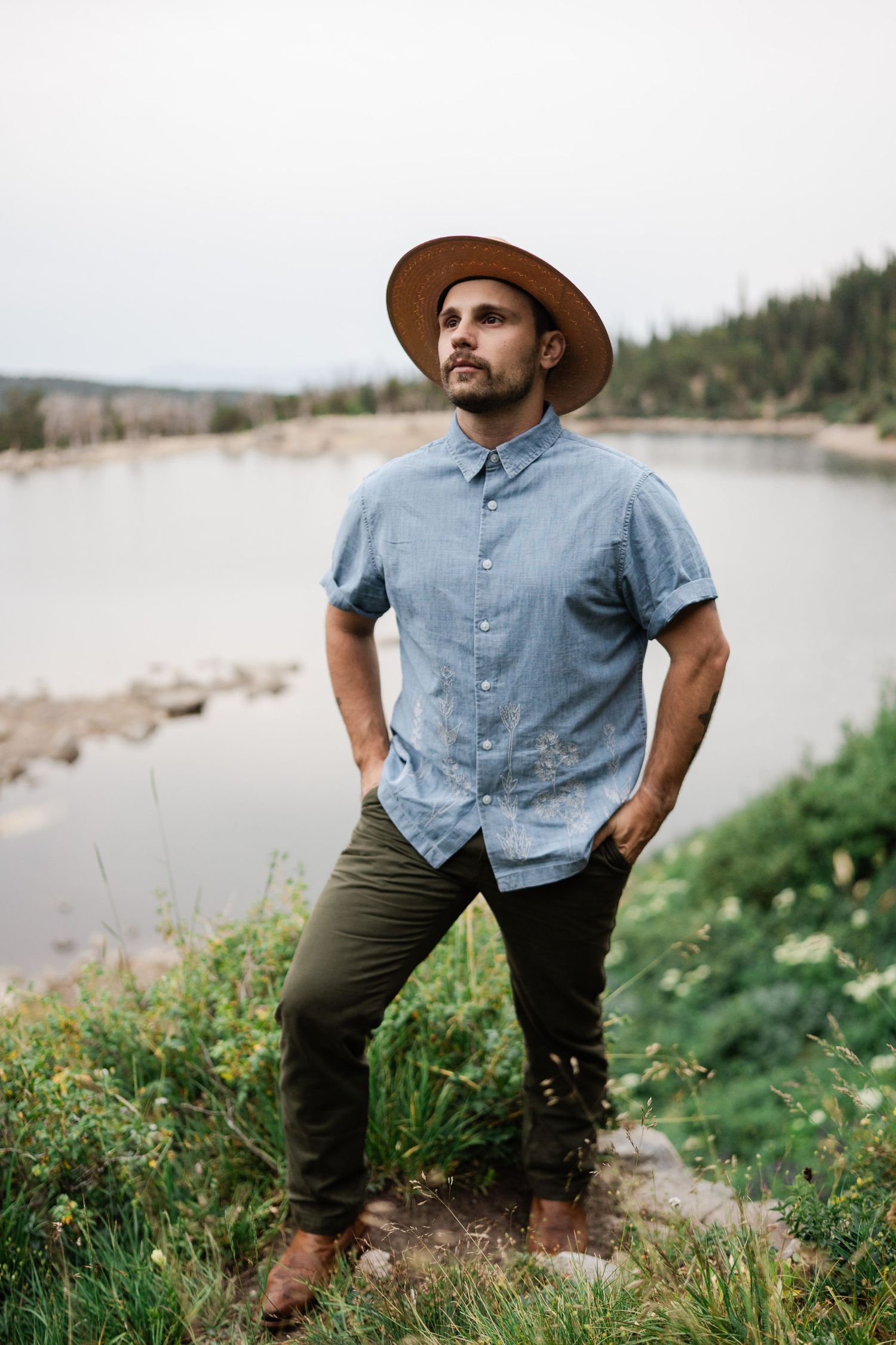 Confident man in tan wide-brim hat and blue shirt standing by a mountain lake with hands in pockets.