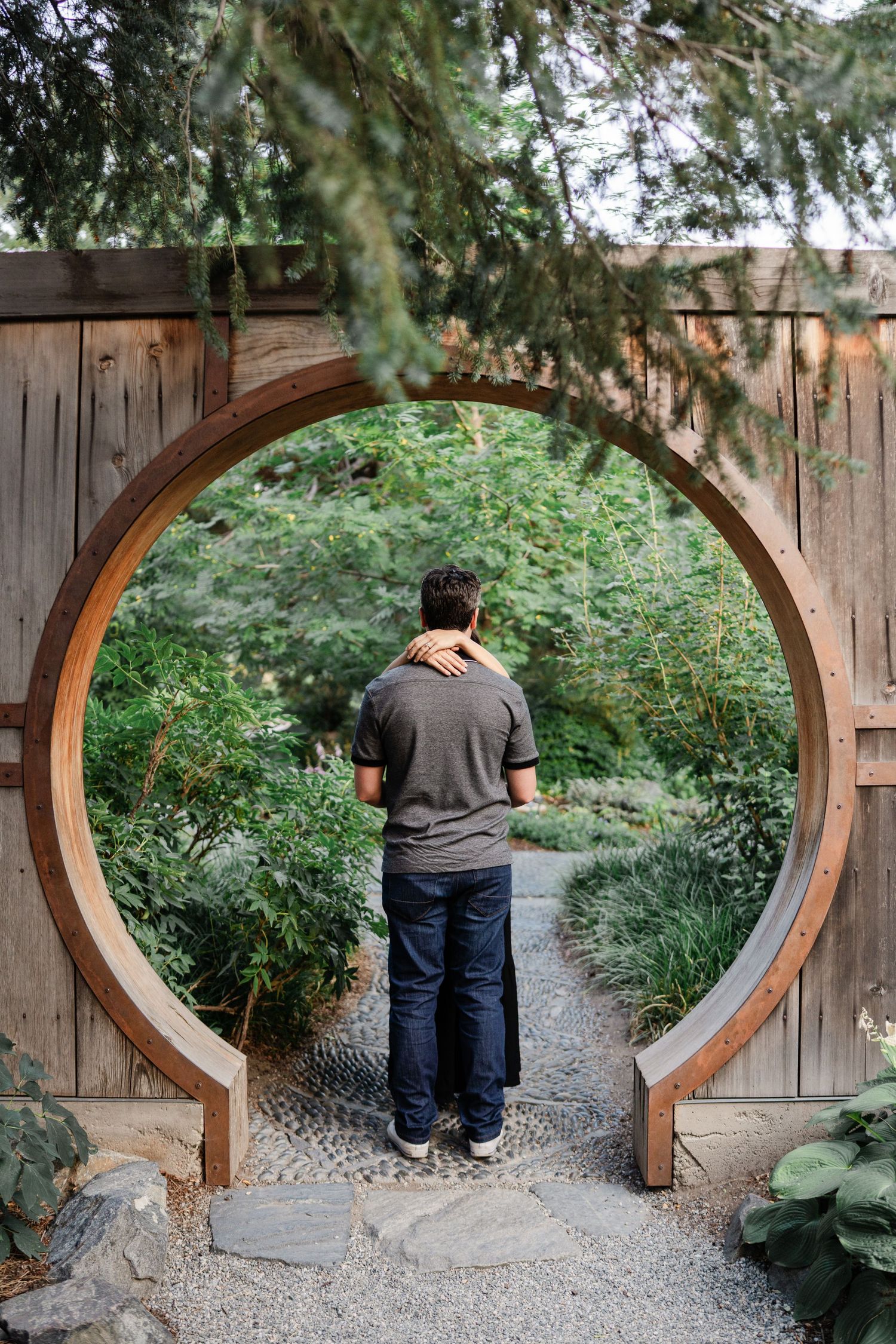 Couple embracing in circular wooden moon gate surrounded by lush garden greenery.