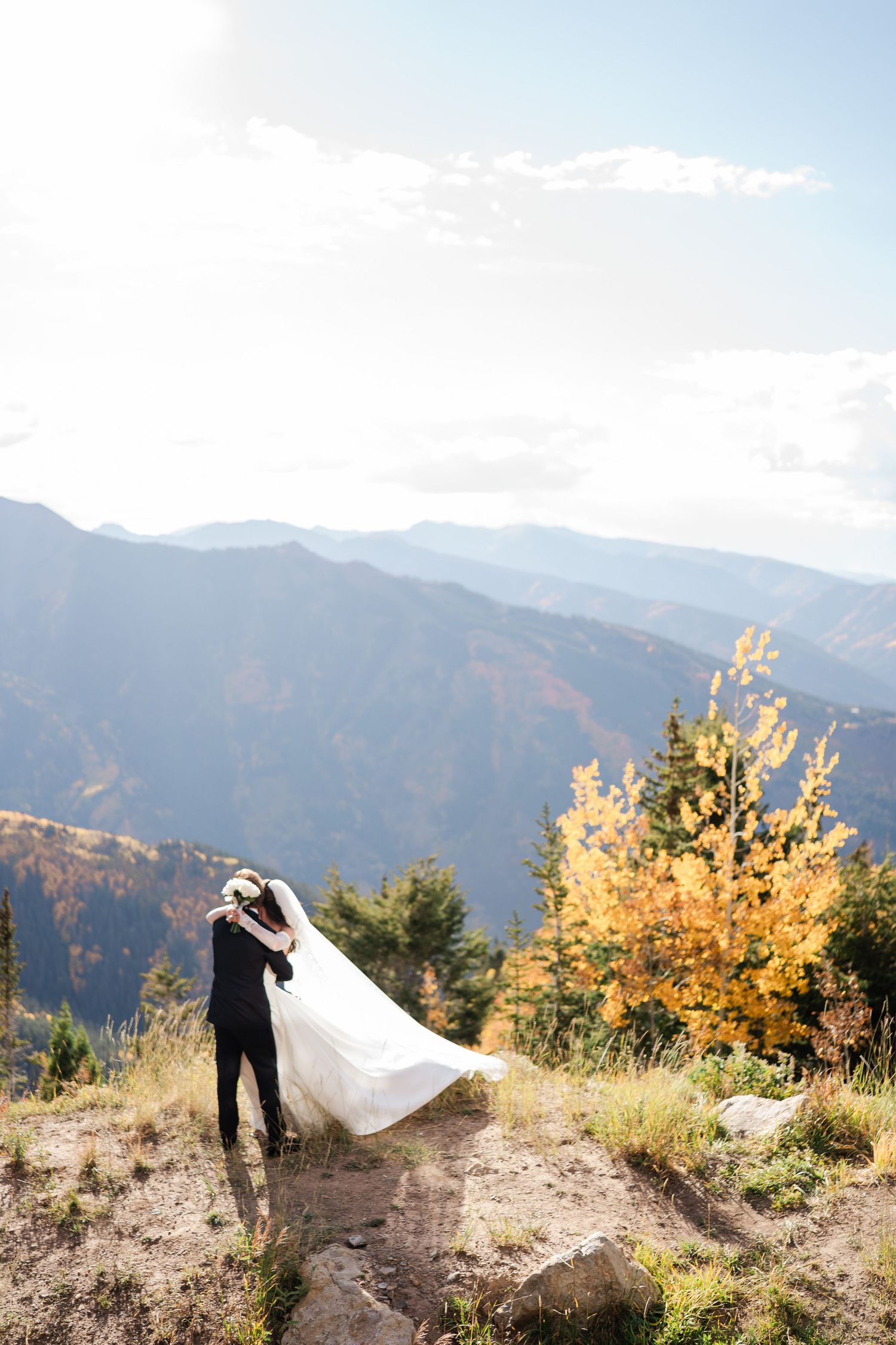Bride and groom embracing on a mountain summit with autumn foliage and sweeping valley views below.