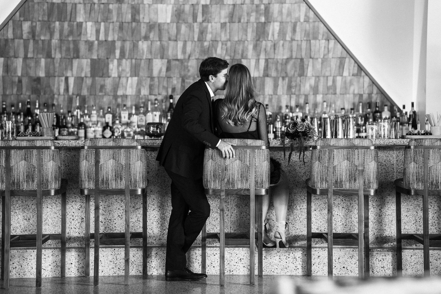 Black and white photo of couple sharing a kiss at an elegant bar with bottle-lined backdrop.