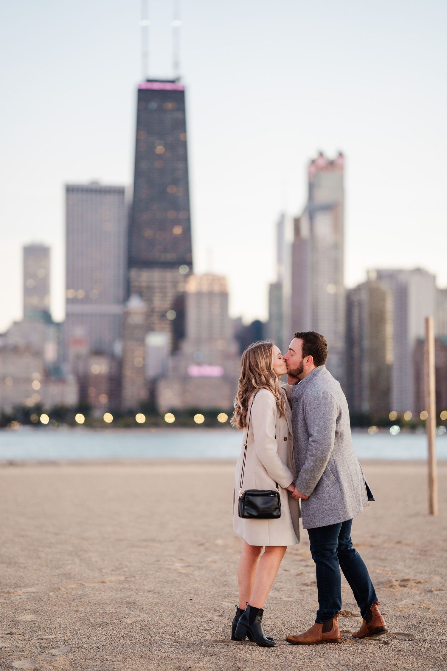 Engaged couple sharing a kiss on Chicago beach with iconic John Hancock skyscraper skyline at golden hour.