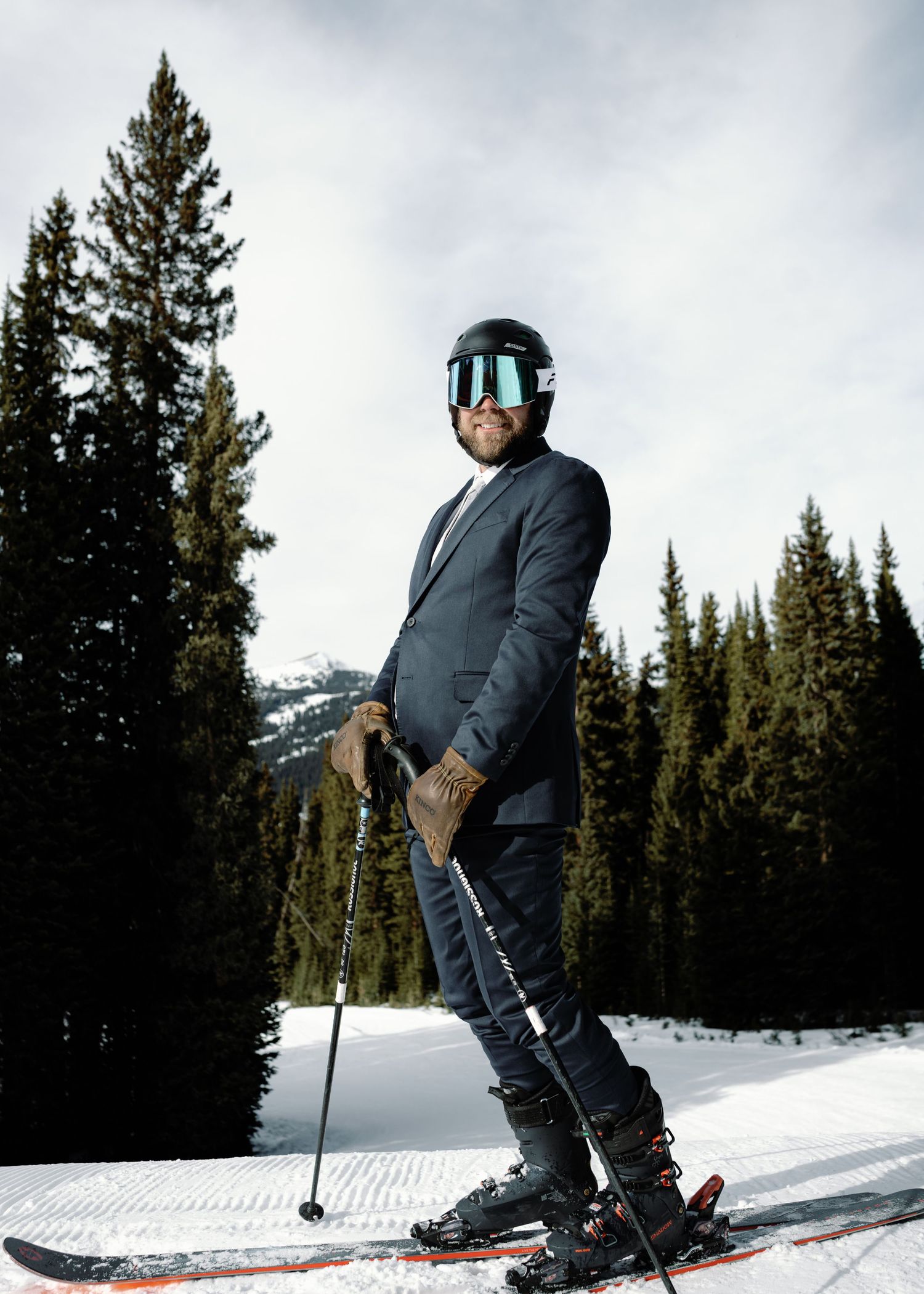 Man in navy suit and teal ski goggles poses confidently on skis against snowy mountain backdrop.