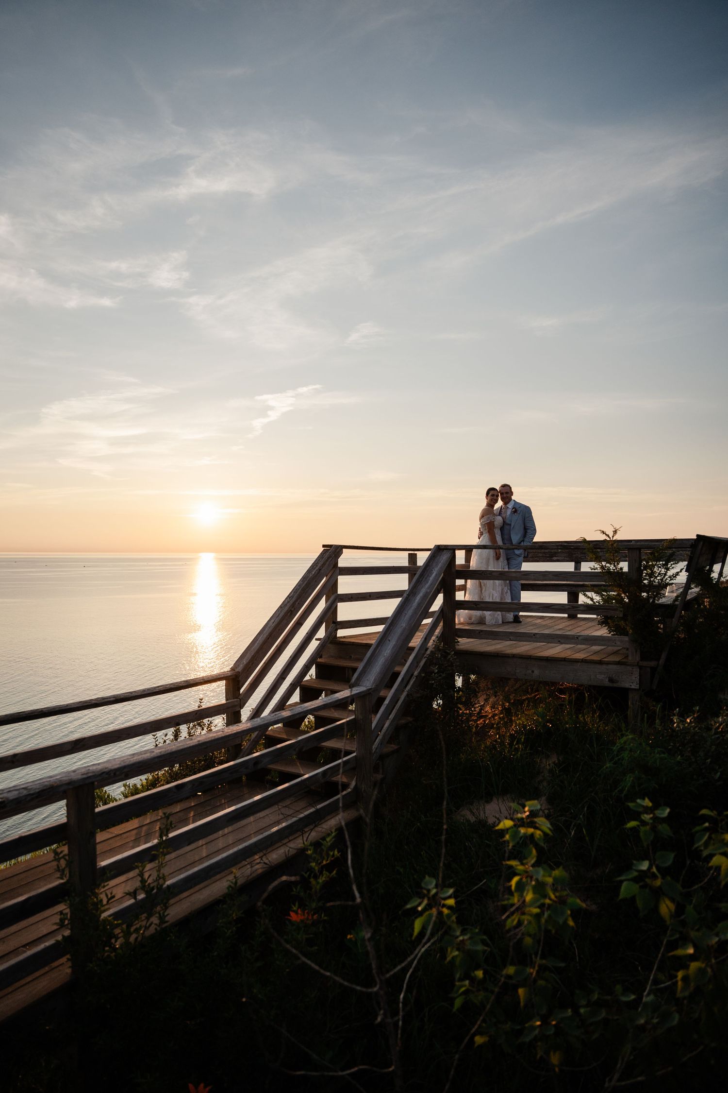 Newlyweds share a romantic moment on wooden overlook deck as sun sets over calm ocean waters.