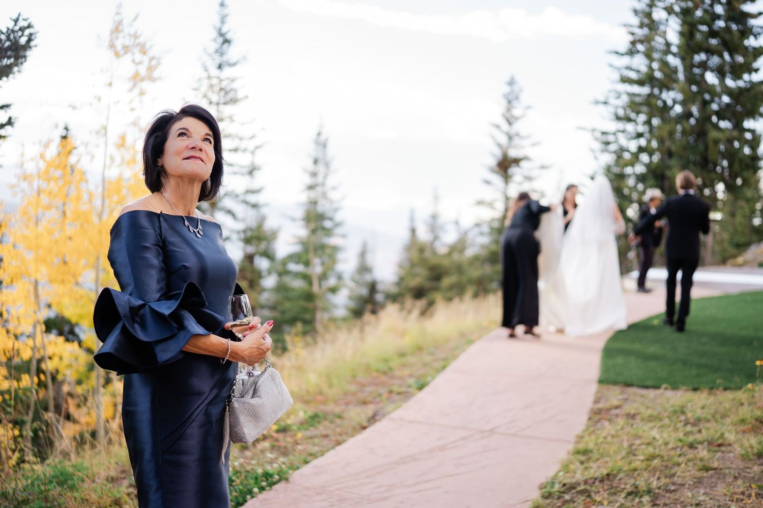 Elegant woman in navy dress watches wedding ceremony from a tree-lined garden path at an outdoor mountain venue.