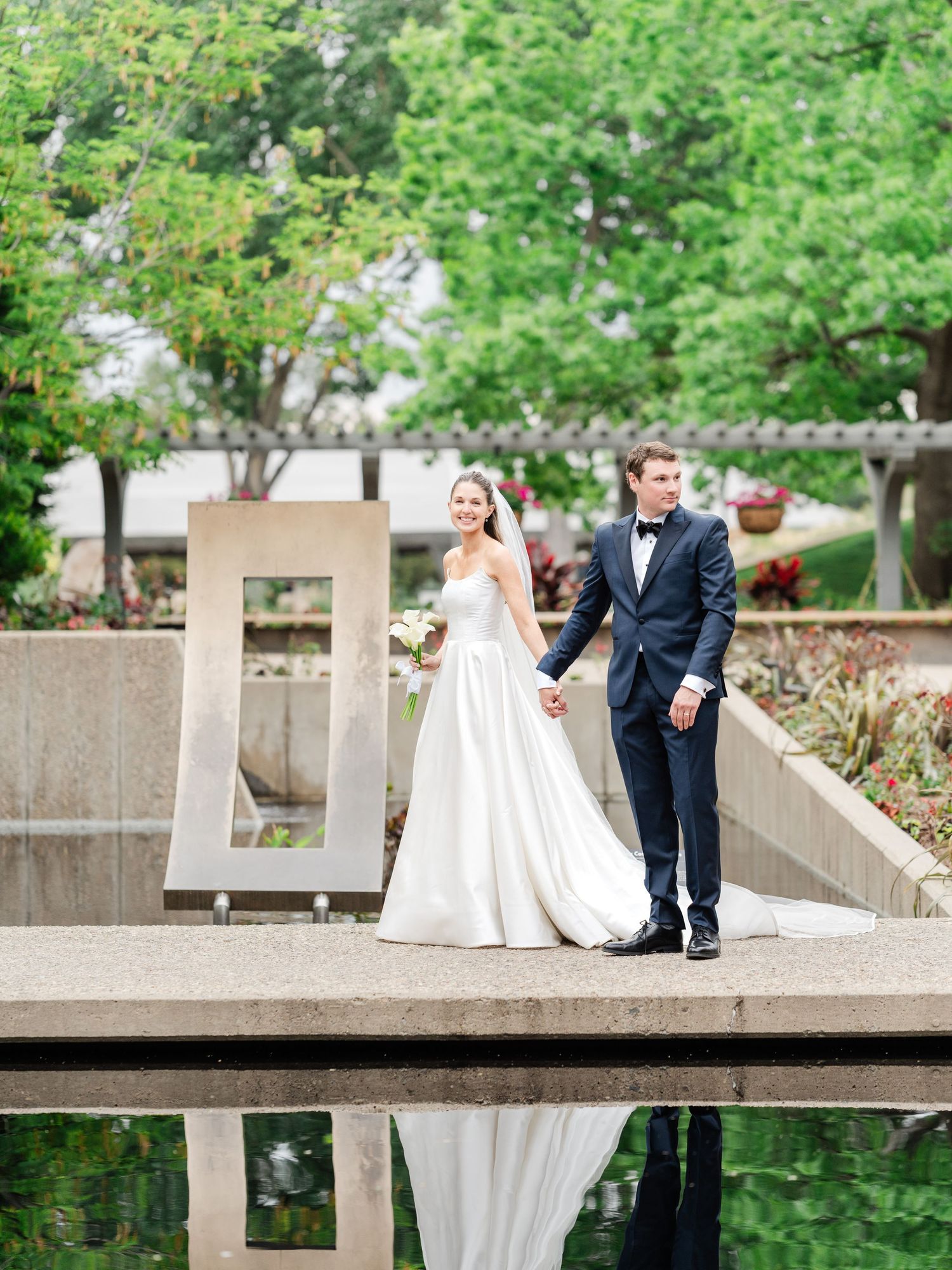 Bride and groom hold hands beside a modern sculpture reflected in a garden pond at their elegant wedding.