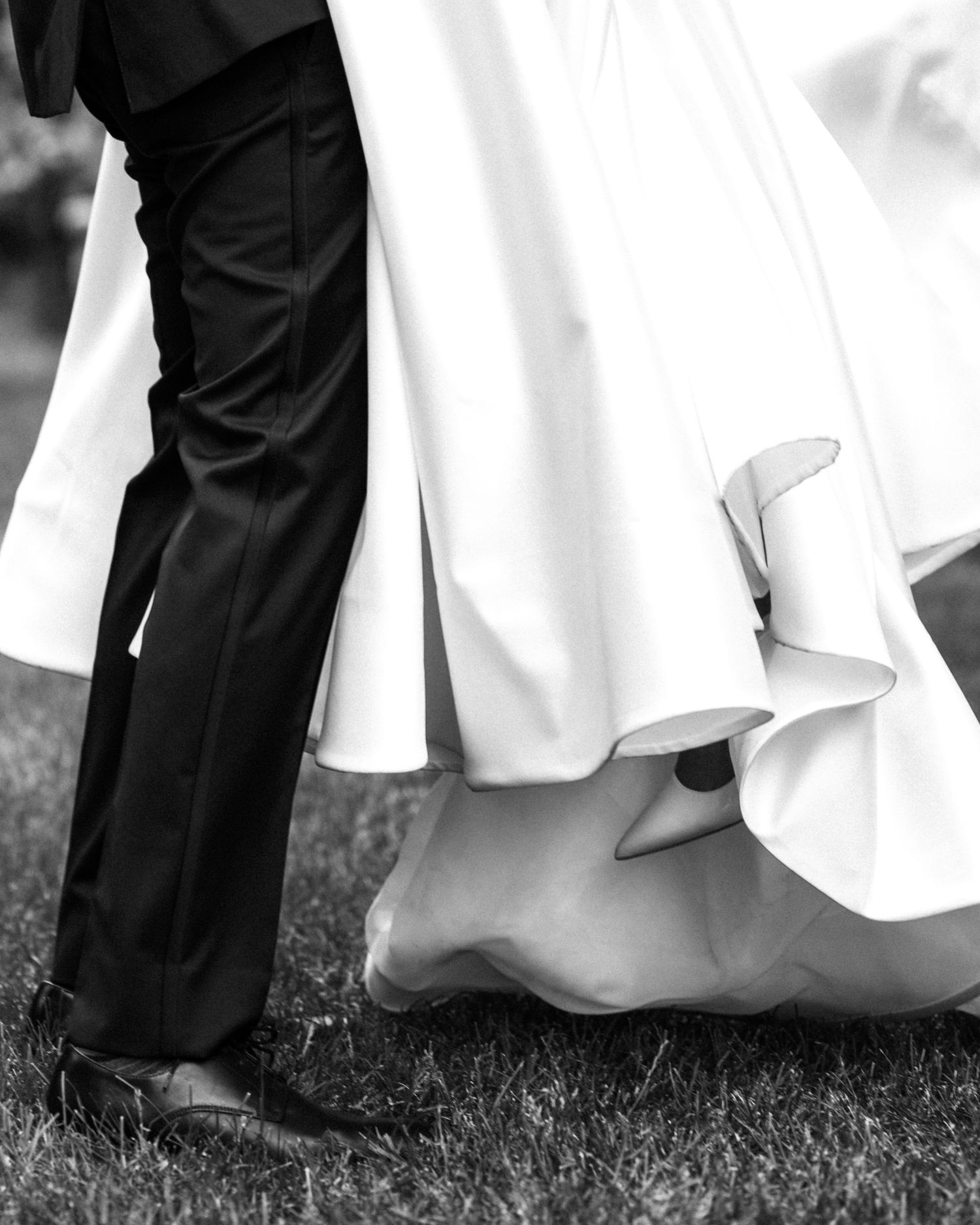 Black and white close-up of bride and groom's feet, showcasing flowing white wedding gown and classic black dress shoes.