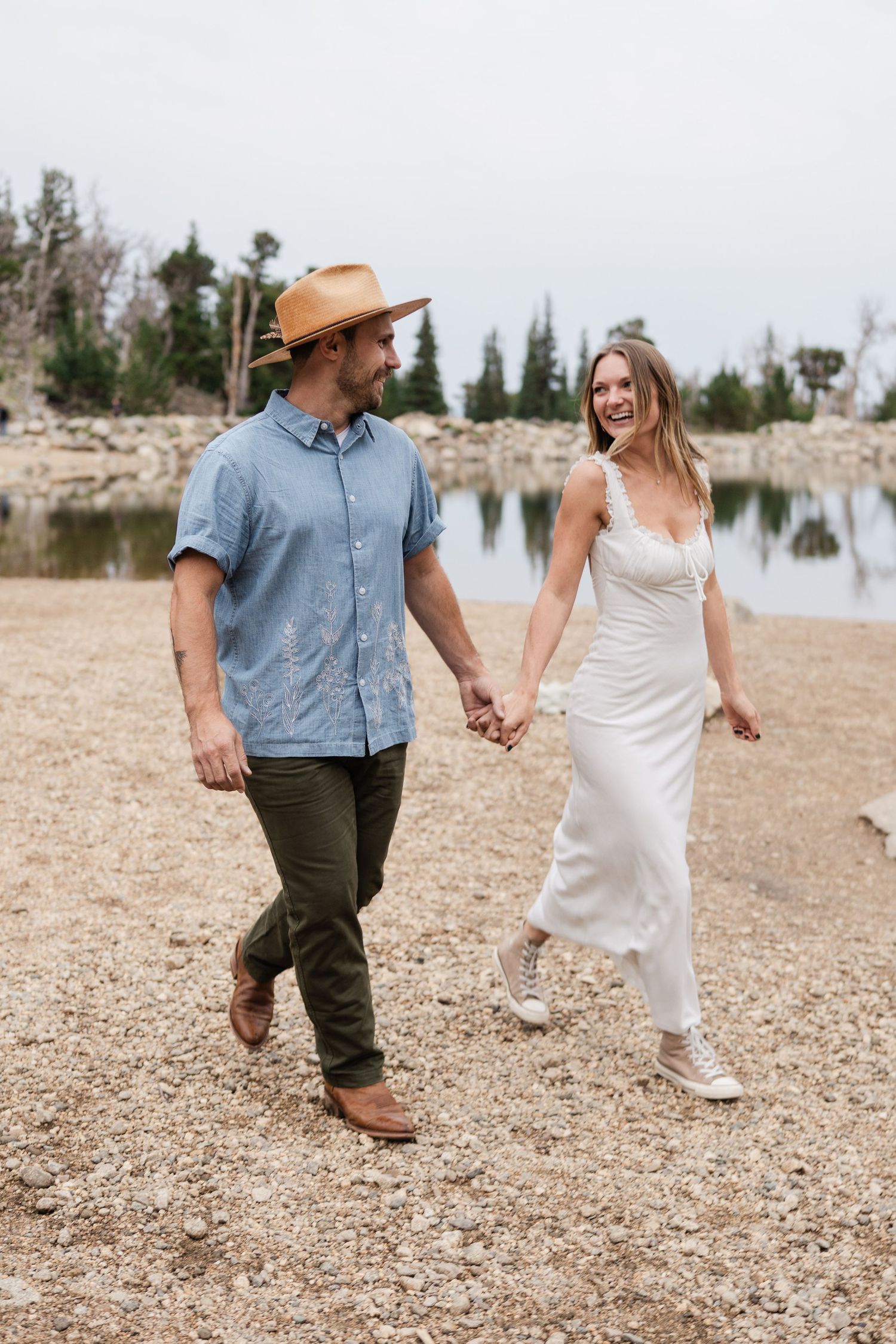 Happy couple holding hands and walking along a rocky lakeside, woman in white dress and man in blue shirt and tan hat.