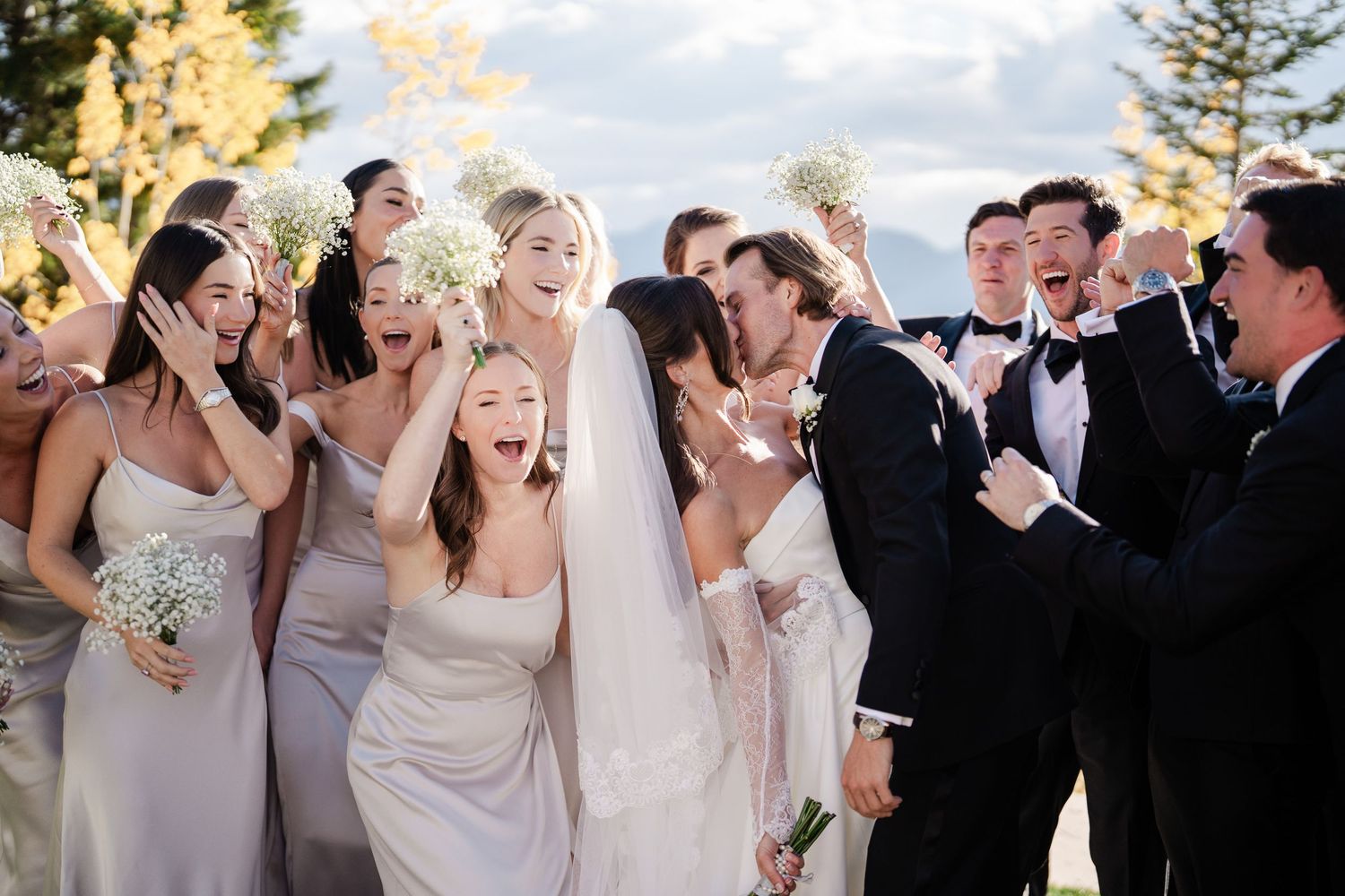 Joyful wedding party celebrating with bride and groom surrounded by cheering bridesmaids and groomsmen outdoors.