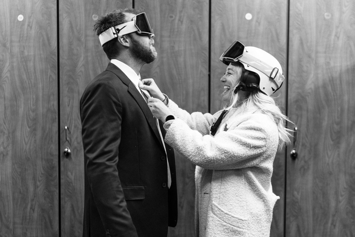 Couple wearing ski goggles adjusting each other's attire in a black and white portrait.