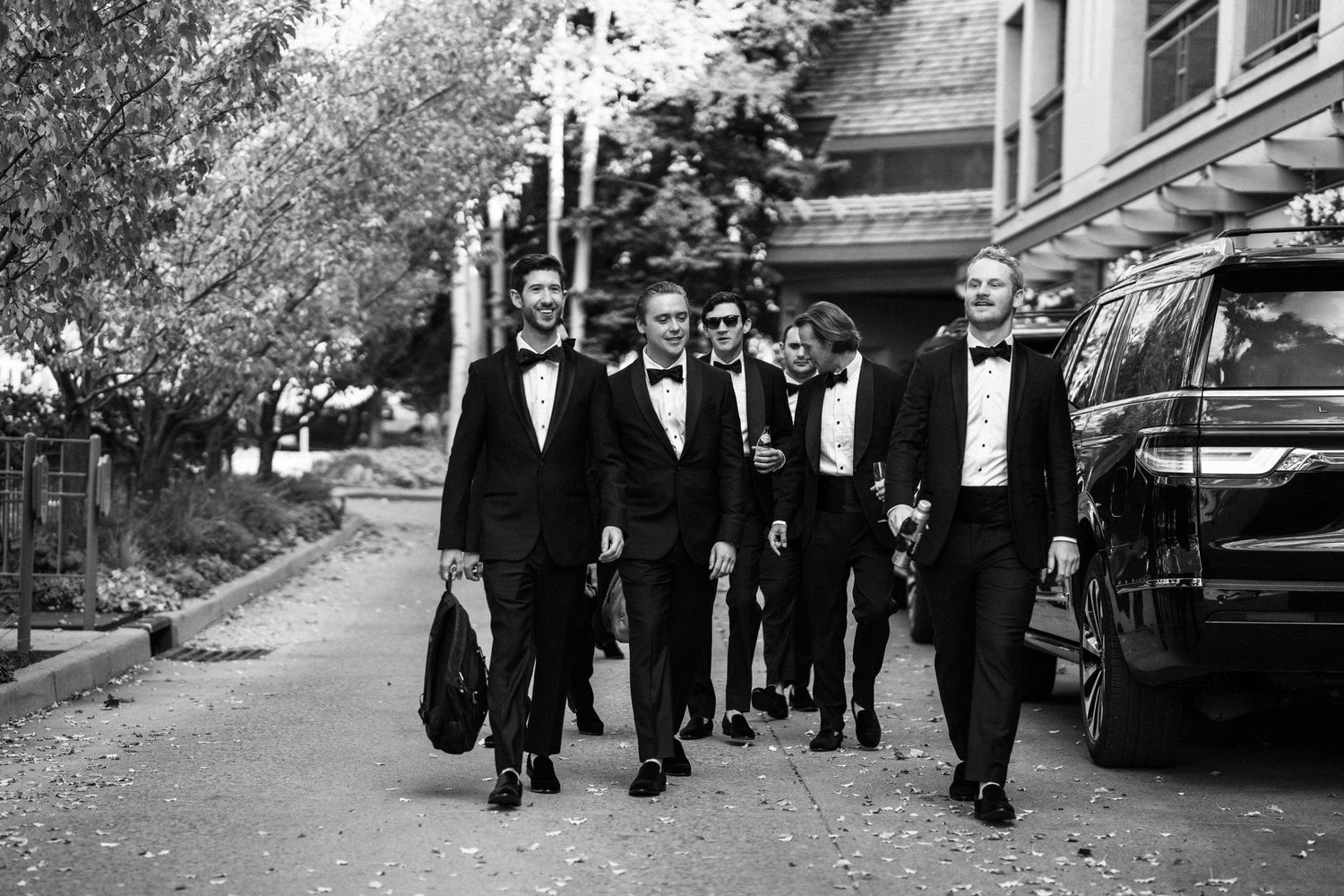 Black and white photo of groomsmen in tuxedos walking confidently along a tree-lined path.