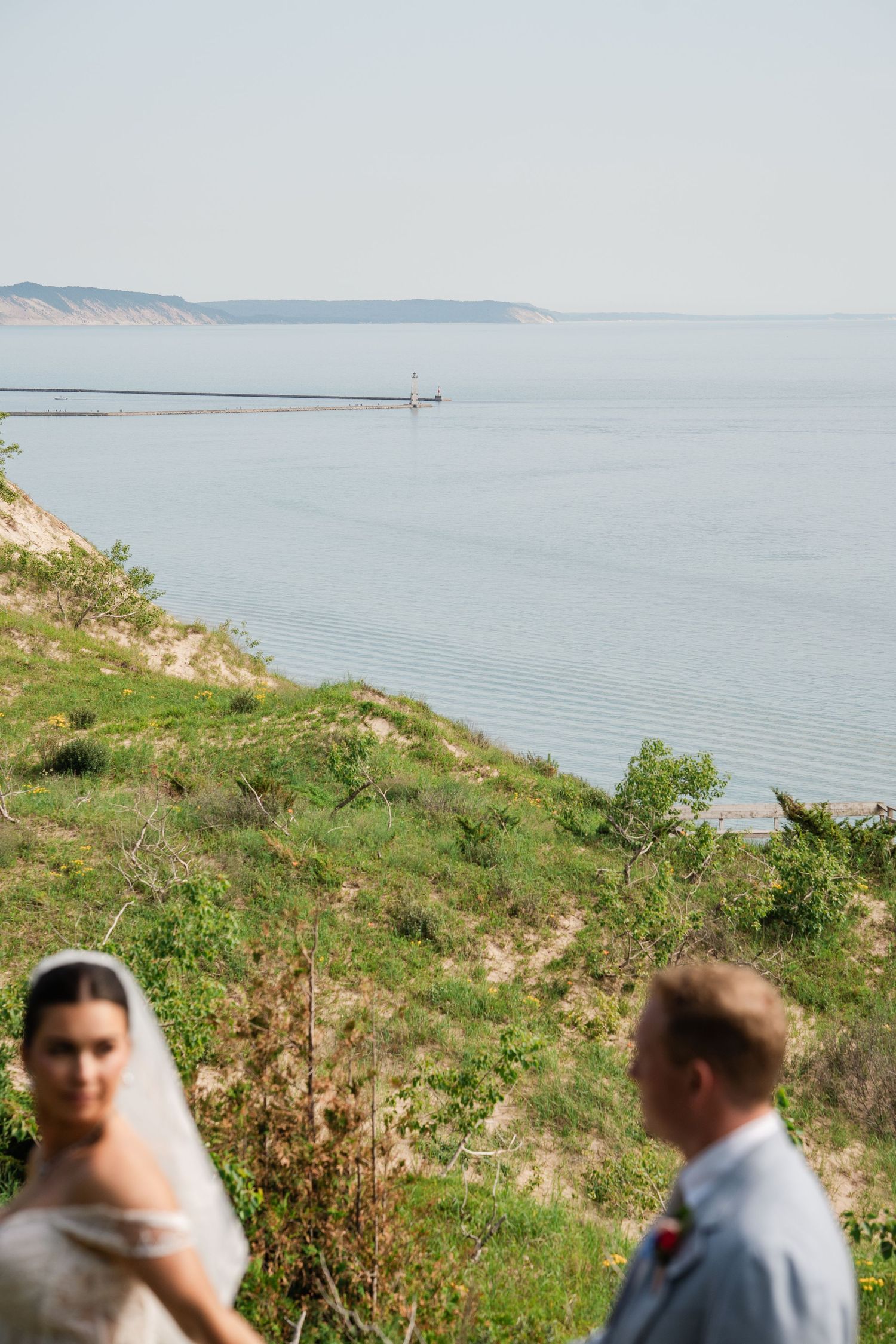 Bride in lace veil and groom standing on a grassy coastal bluff overlooking a calm blue lake.