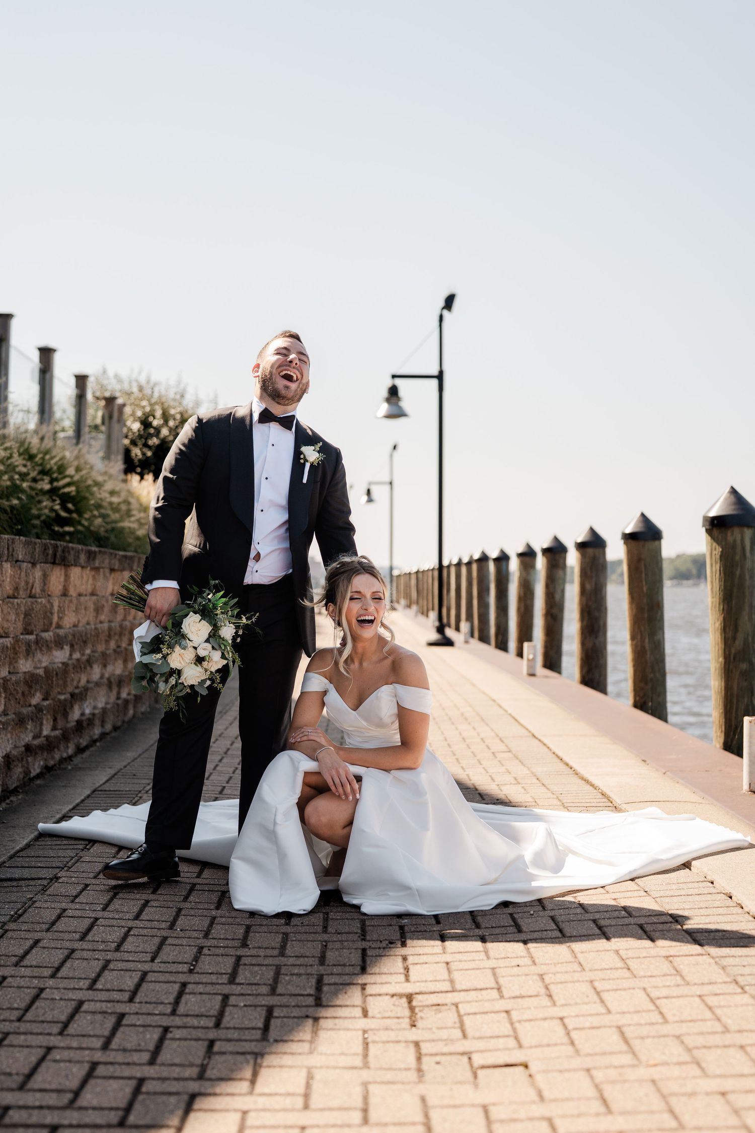 Laughing bride and groom posing on a waterfront brick walkway on a sunny wedding day.