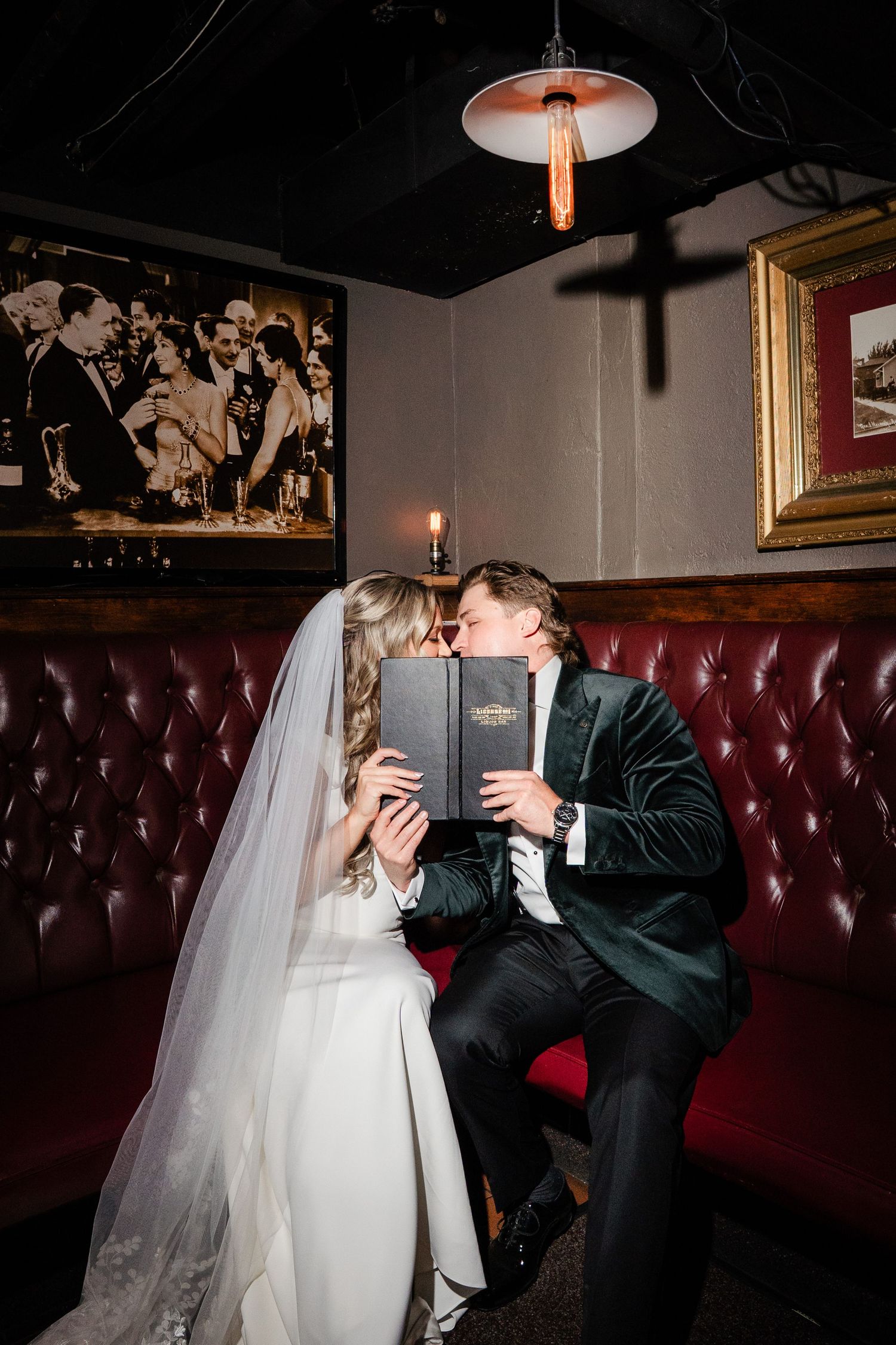 Bride and groom sharing a kiss behind a menu book in a cozy red leather booth at a bar.