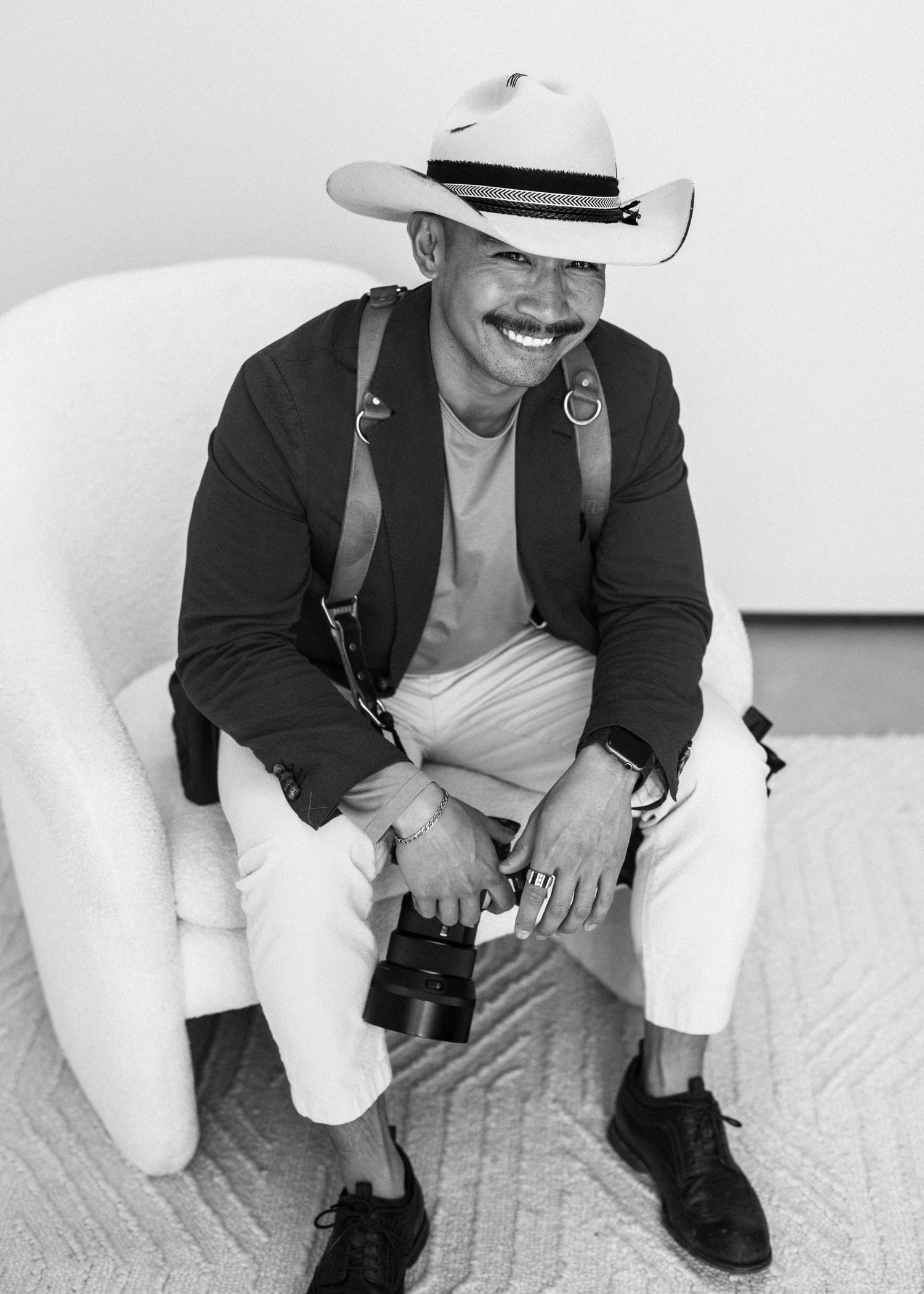Stylish wedding photographer in cowboy hat sits with camera in black and white portrait.
