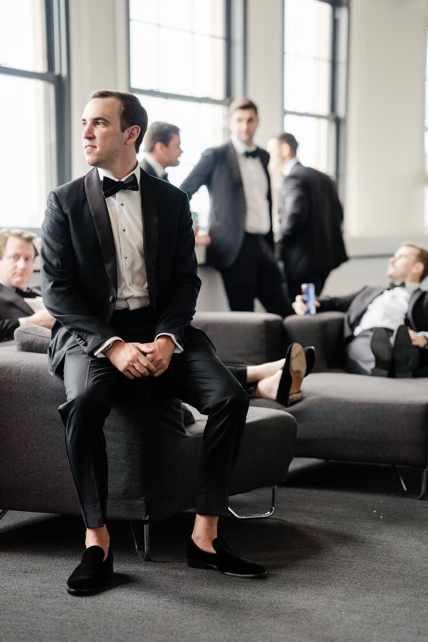 Groom in black tuxedo with bow tie sits confidently while groomsmen relax in modern lounge before wedding.