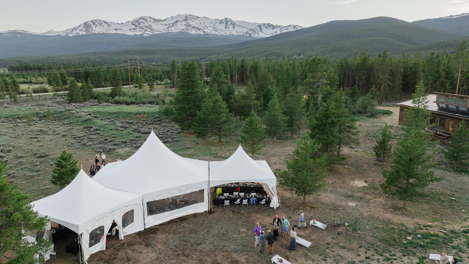 Aerial view of white wedding reception tent surrounded by pine trees with mountain landscape backdrop.