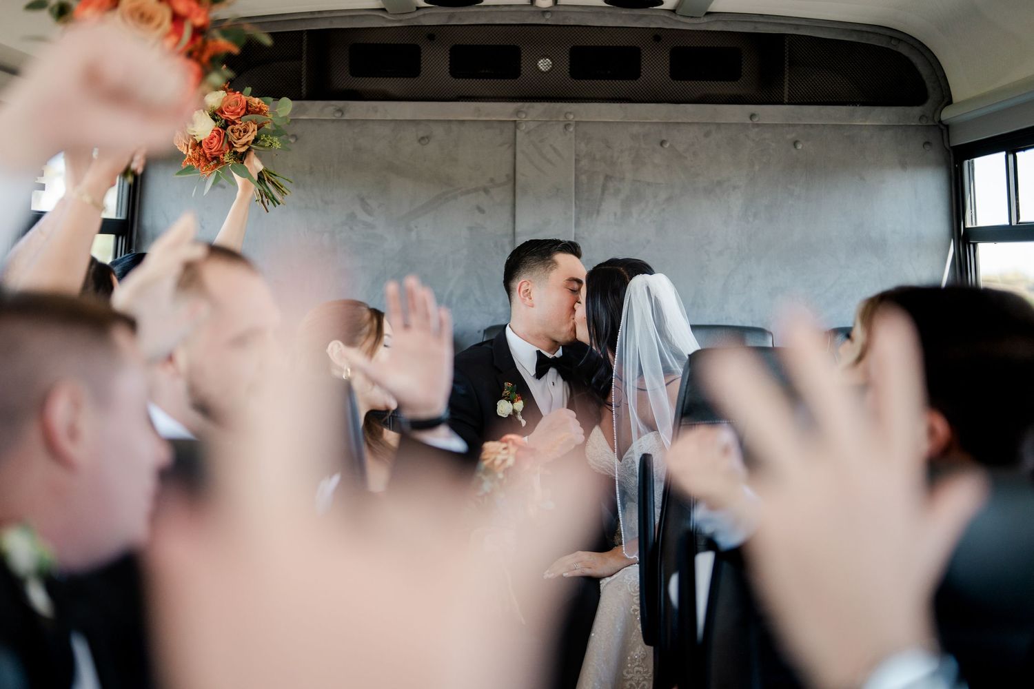 Bride and groom sharing a kiss on party bus surrounded by cheering wedding party guests.