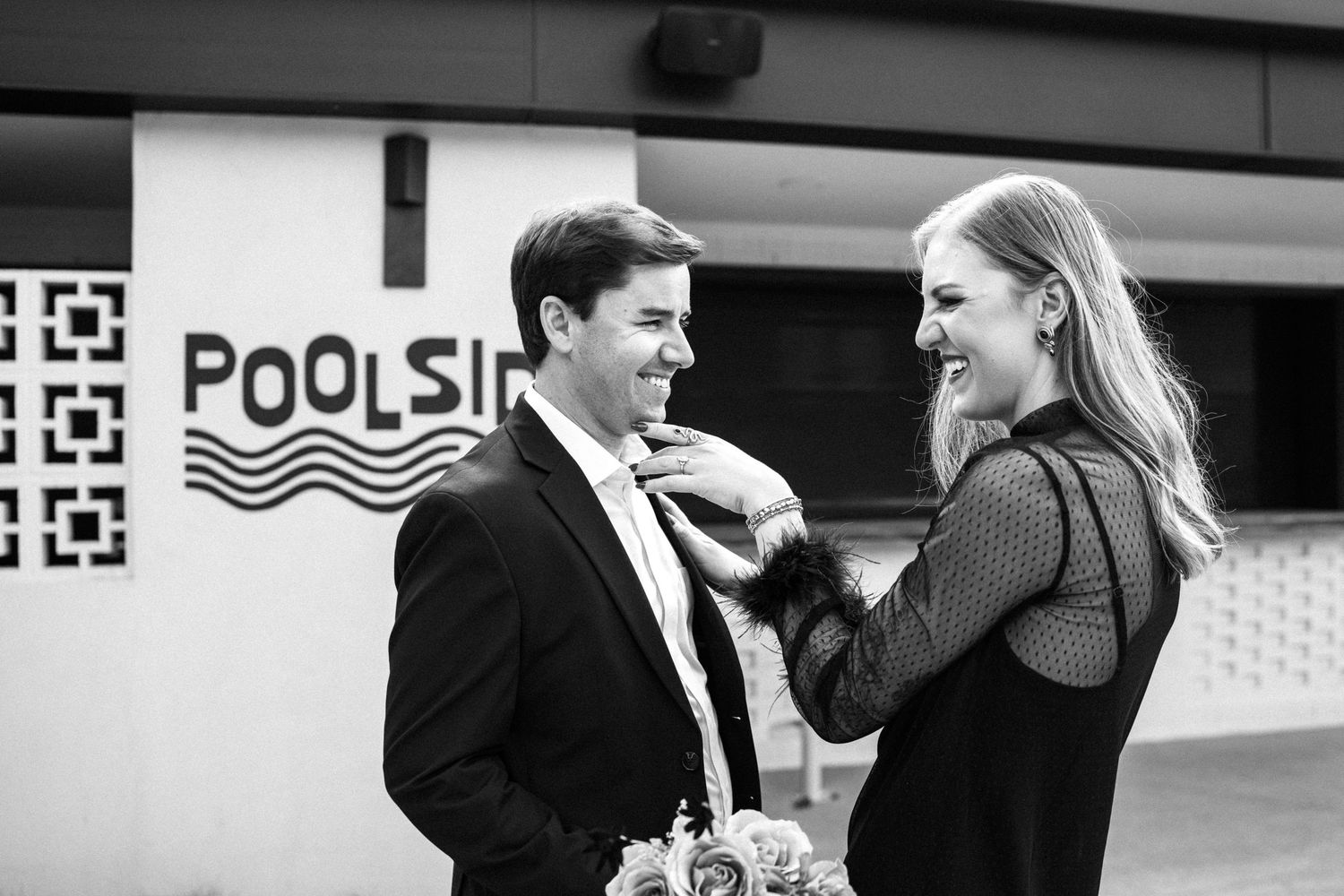 Black and white photo of smiling couple near poolside sign, woman adjusting mans tie outdoors.