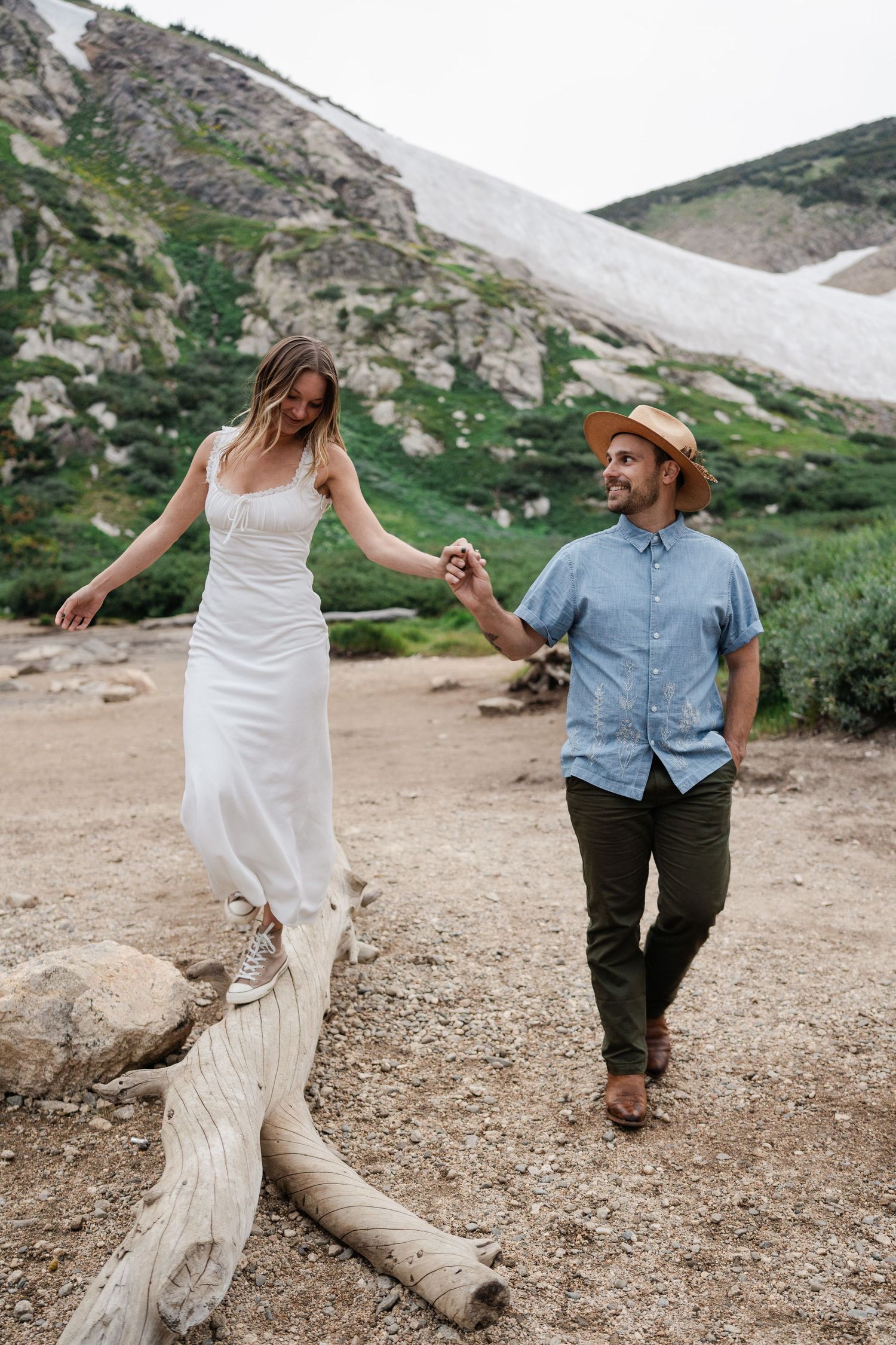 A smiling couple holds hands while walking near a fallen log with snow-capped mountains and greenery behind them.