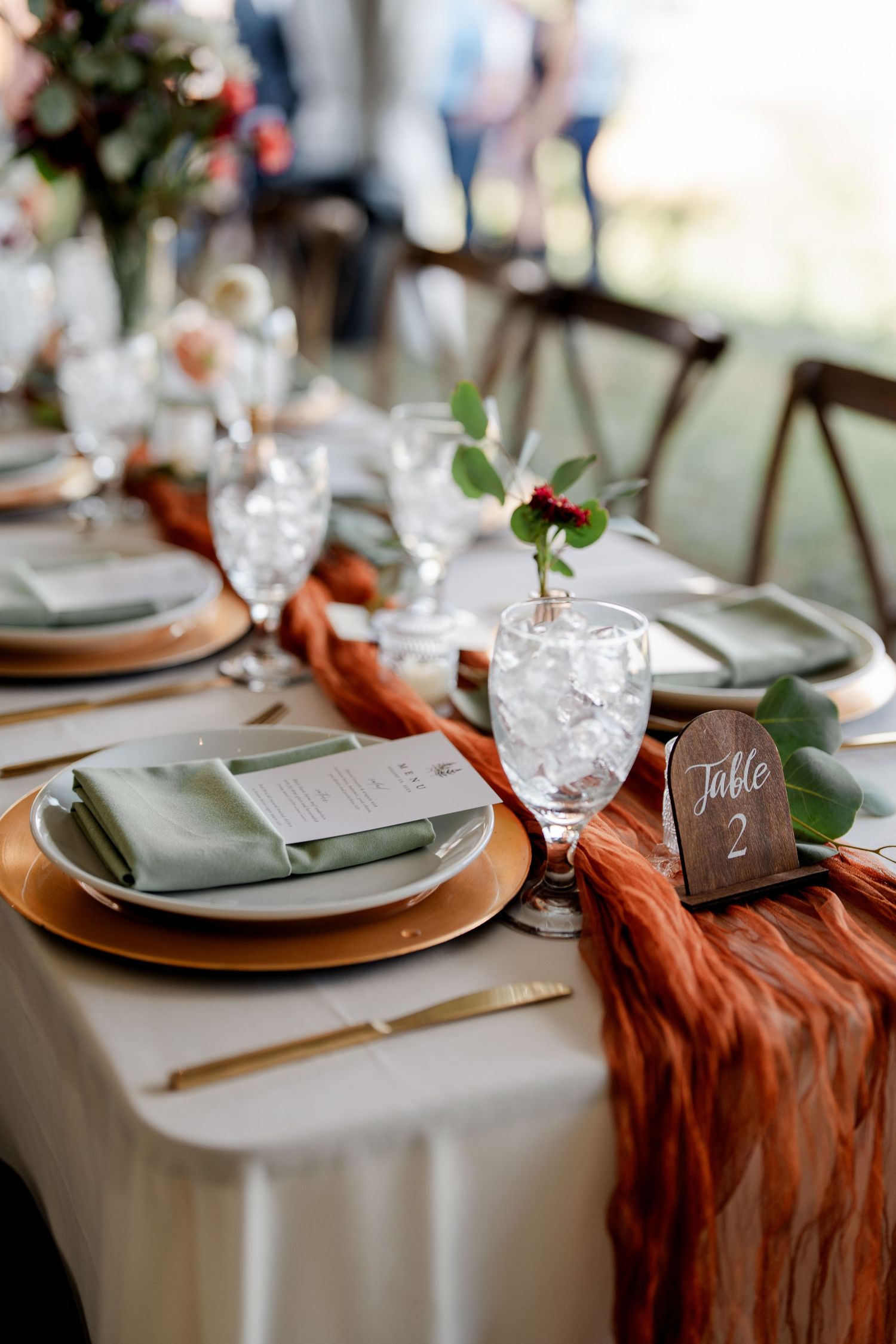 Elegant wedding reception table with rust chiffon runner, gold chargers, sage napkins, and floral.