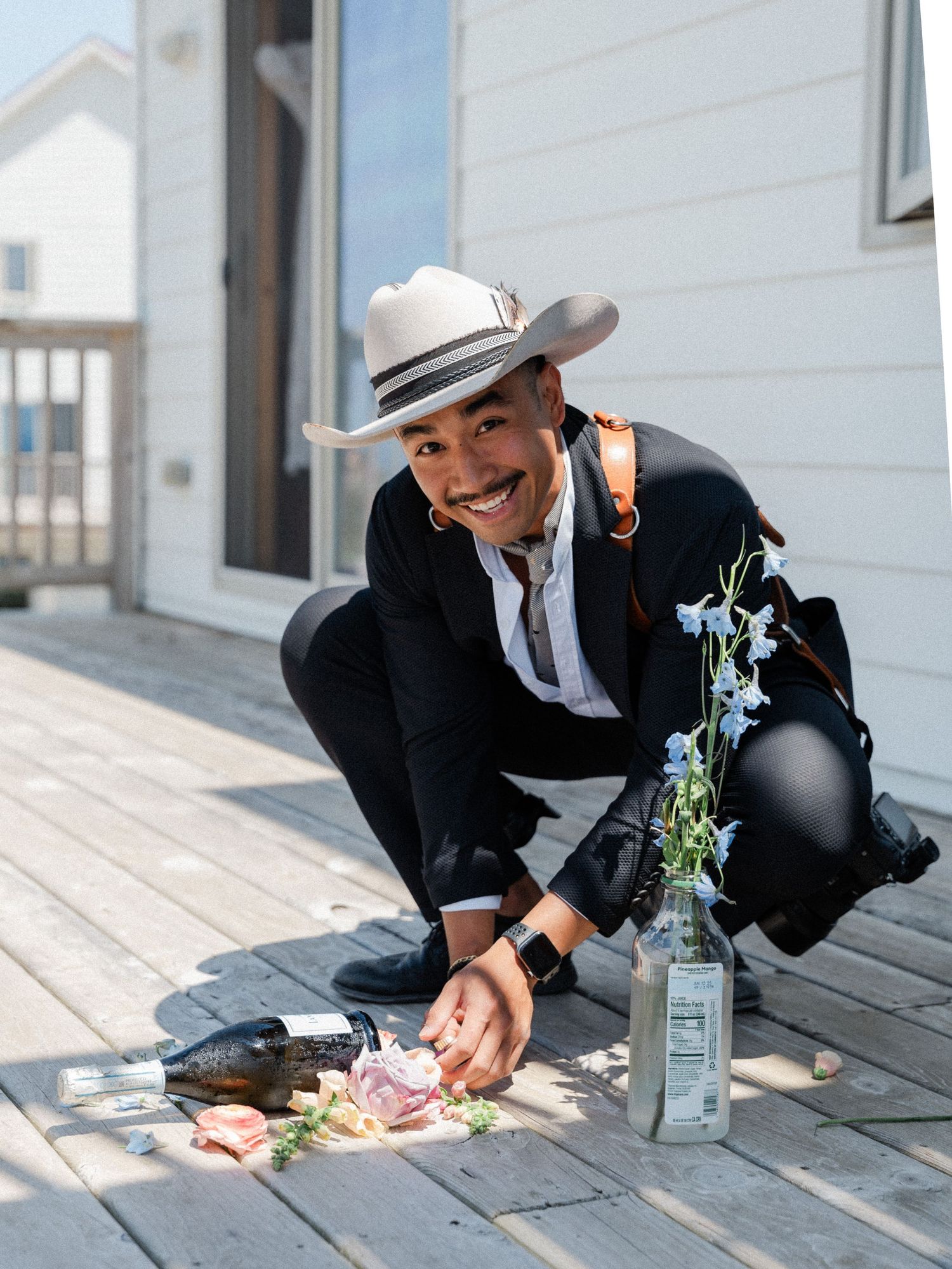 Smiling man in cowboy hat and black suit arranging floral details on a sunny wooden deck.