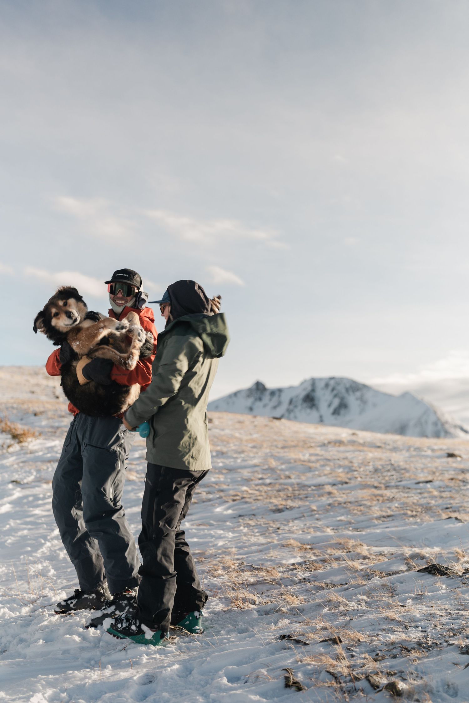 A skier in red jacket holds a dog while a partner in green jacket photographs them on a snowy mountain summit.