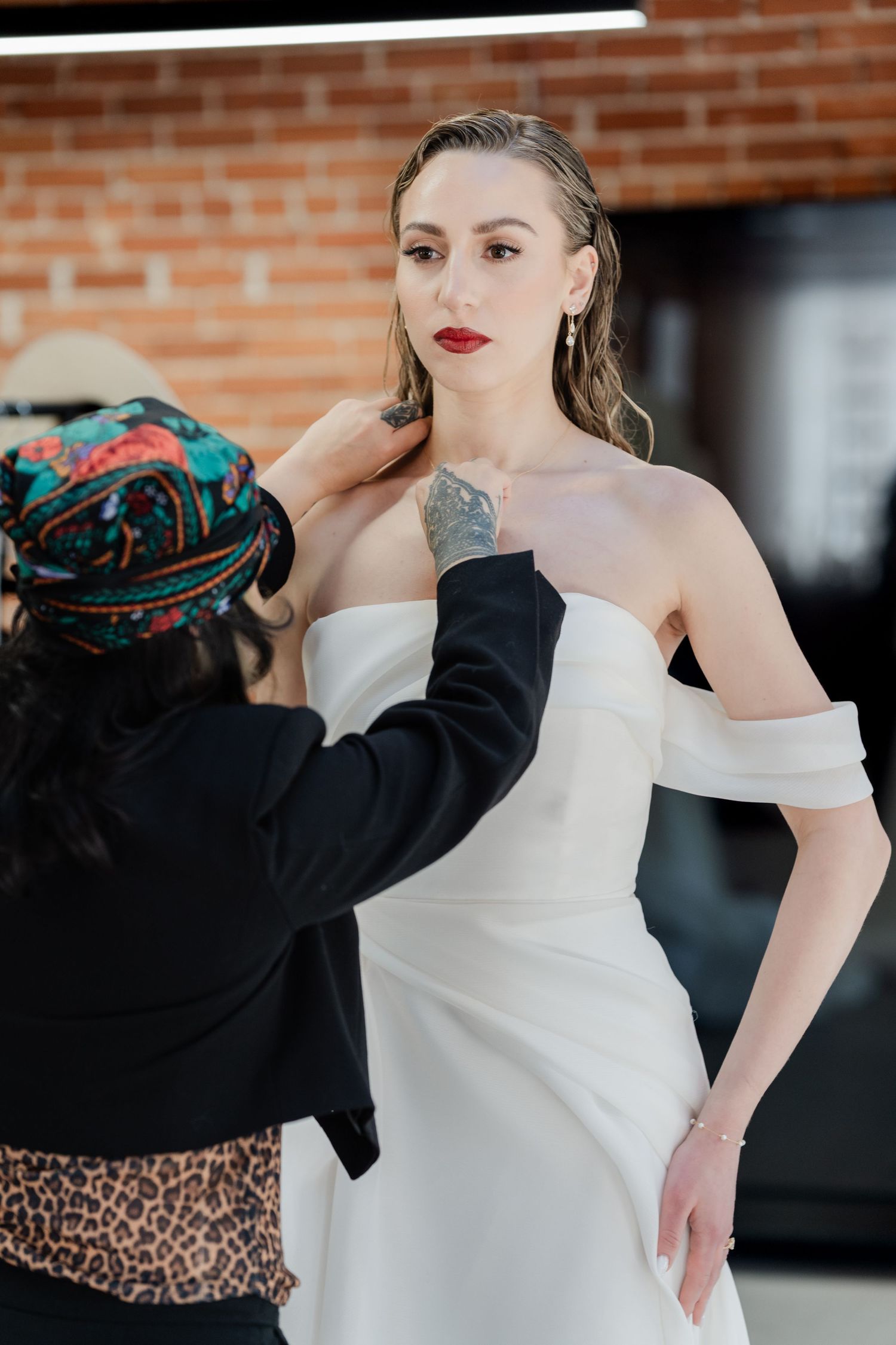 A bridal stylist adjusts a white off-shoulder wedding gown on a model with red lips and a tattoo in a studio.