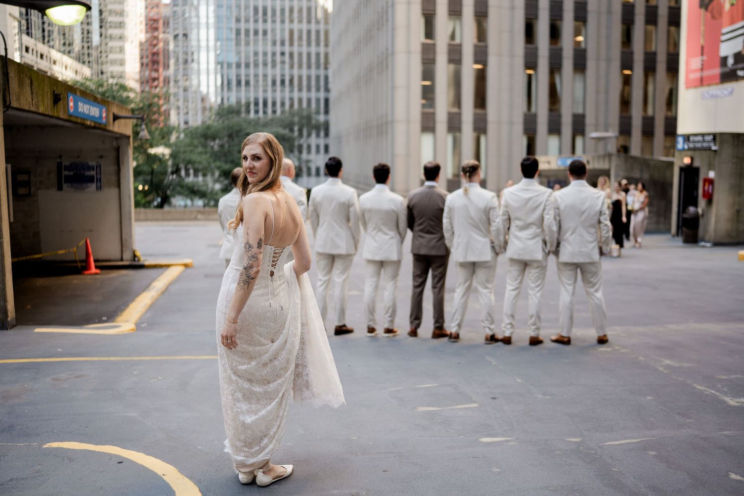 Bride in lace gown looking back as groomsmen in white suits stand in a row on a city street.