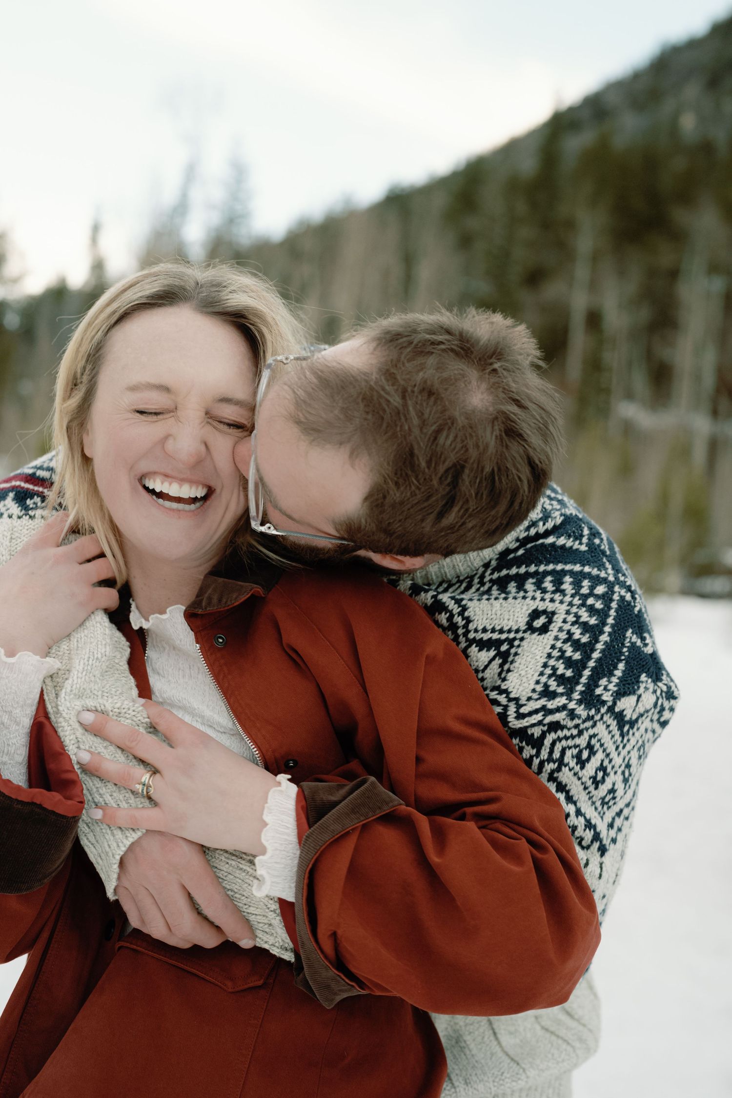 Joyful couple embracing in snow, woman laughing as man kisses her cheek during winter engagement session.