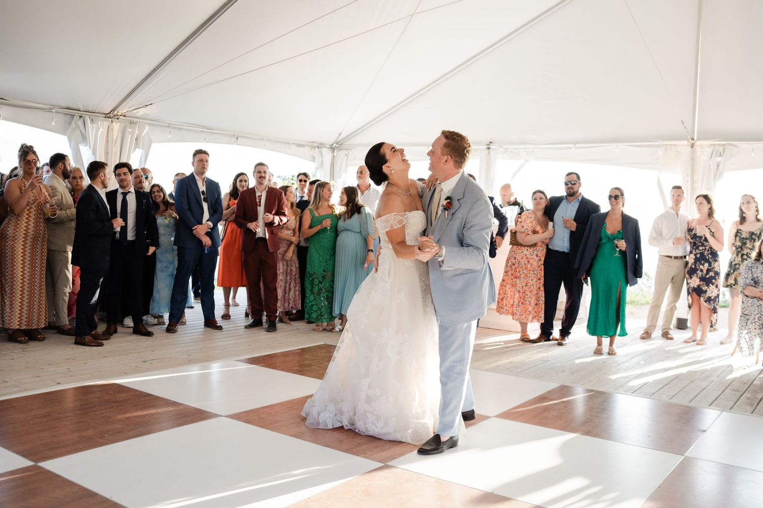 Bride and groom share their first dance on a checkered floor surrounded by applauding wedding guests.