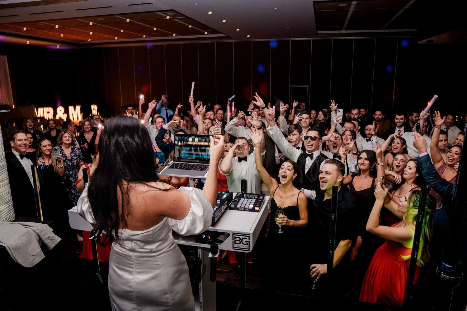 Bride at DJ booth with large wedding party crowd cheering behind her on dance floor.