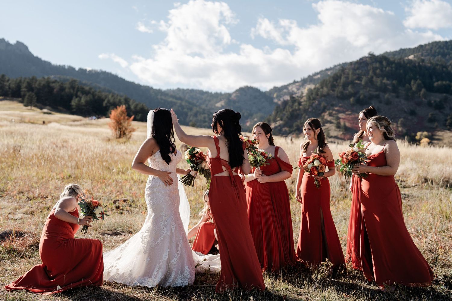 Bride with bridesmaids in red dresses holding bouquets in a scenic mountain meadow setting.