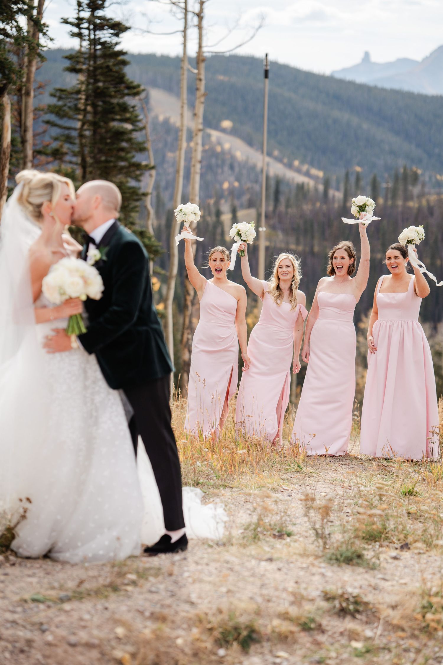 Bride and groom sharing romantic kiss while bridesmaids in pink dresses celebrate with raised bouquets.
