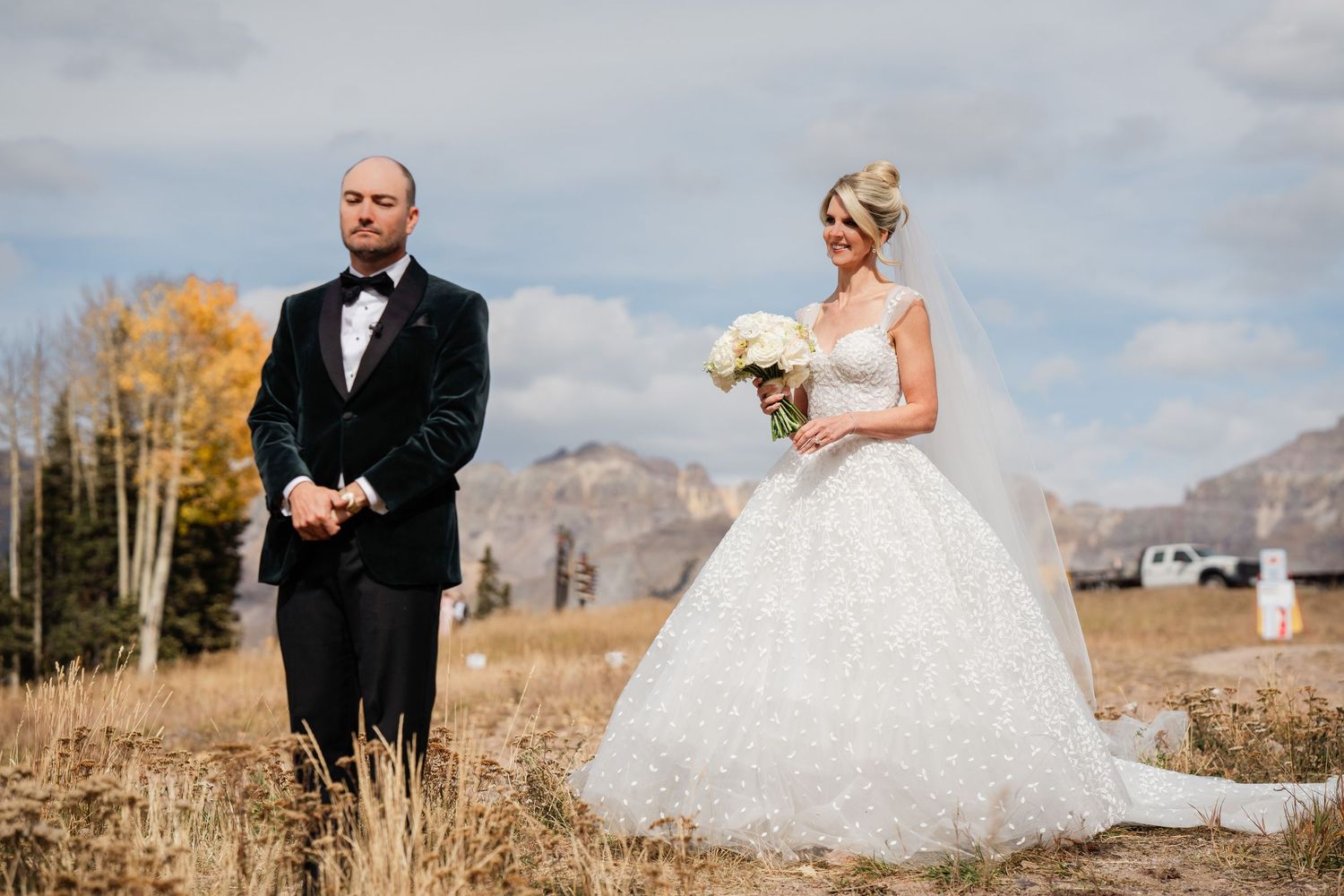 Bride in a ballgown holding white bouquet surprises groom during first look in autumn mountain landscape.
