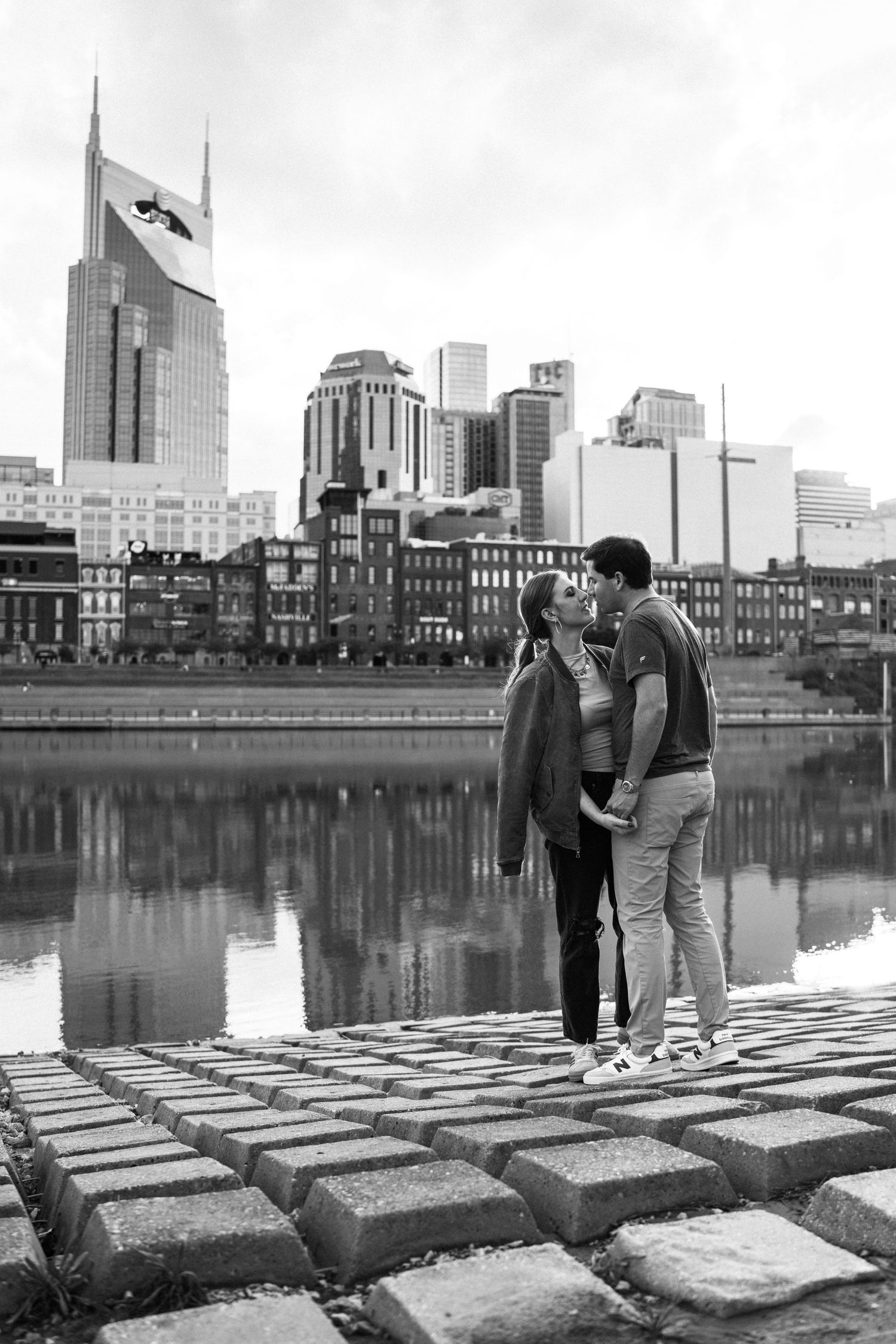 Black and white photo of couple kissing by river with Nashville city skyline reflected in water.