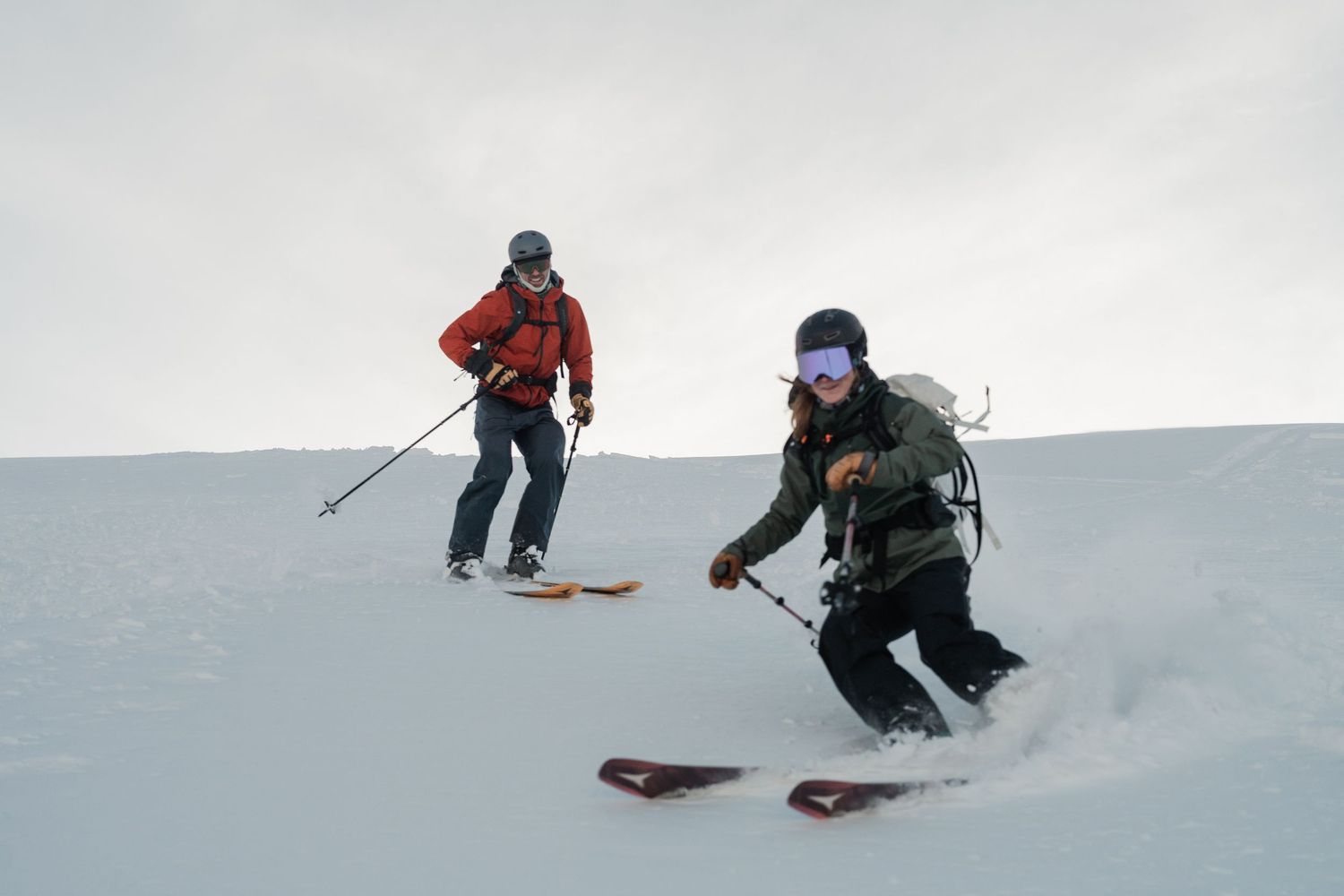Two skiers carving through fresh powder snow on an overcast winter day in the backcountry.