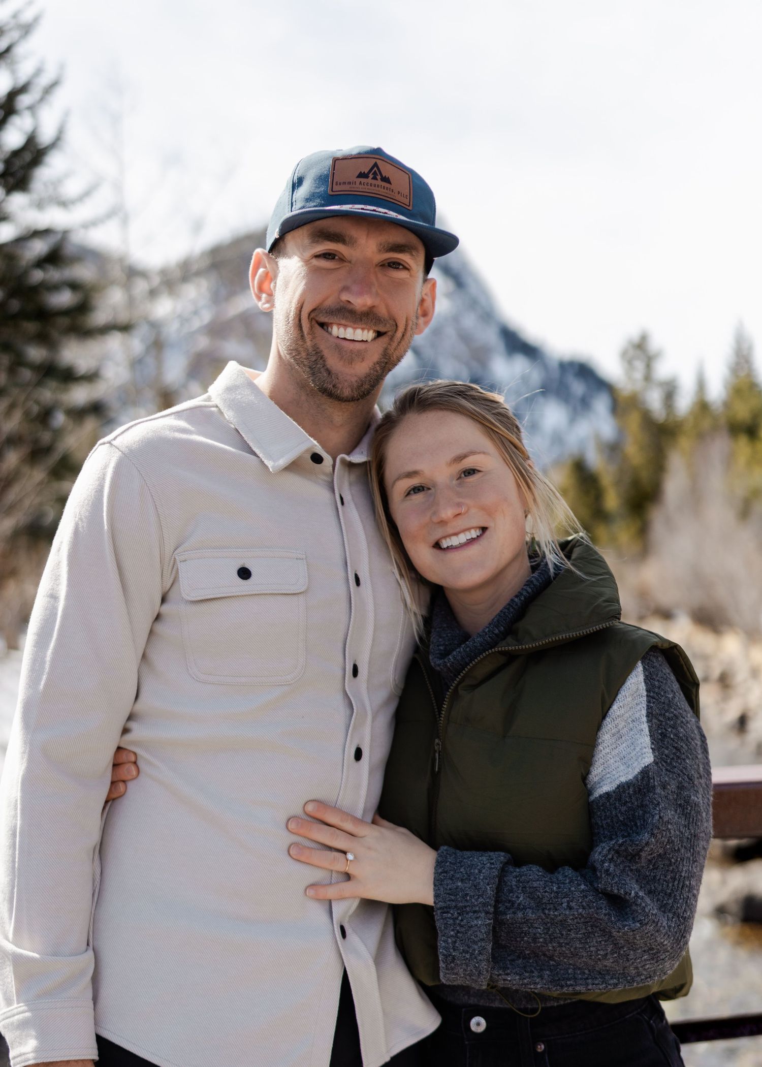 Smiling couple posing outdoors in winter mountain landscape, man wearing cap and white shirt, woman in green vest.