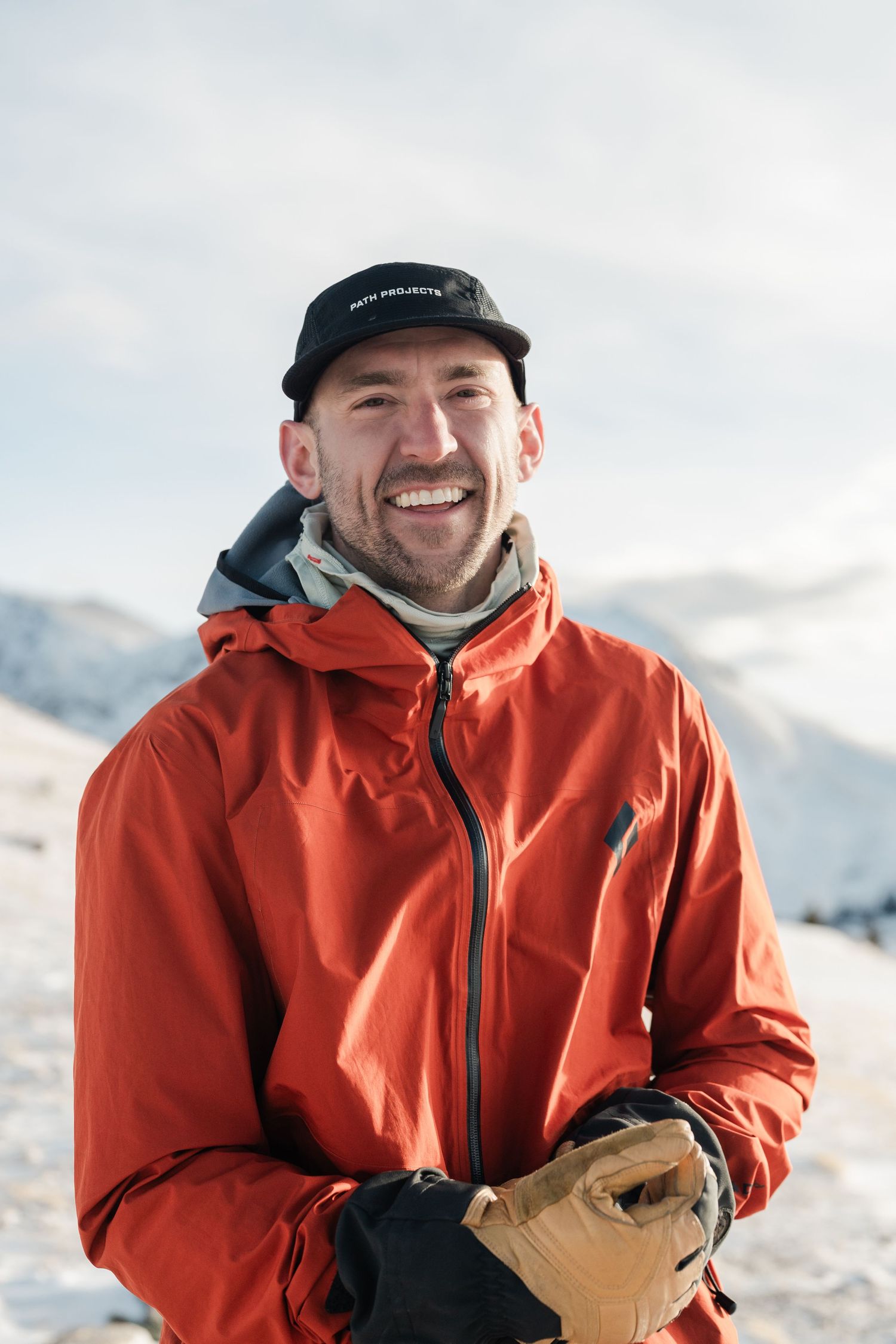 Smiling man in red jacket and black cap standing in snowy mountain landscape on a sunny winter day.