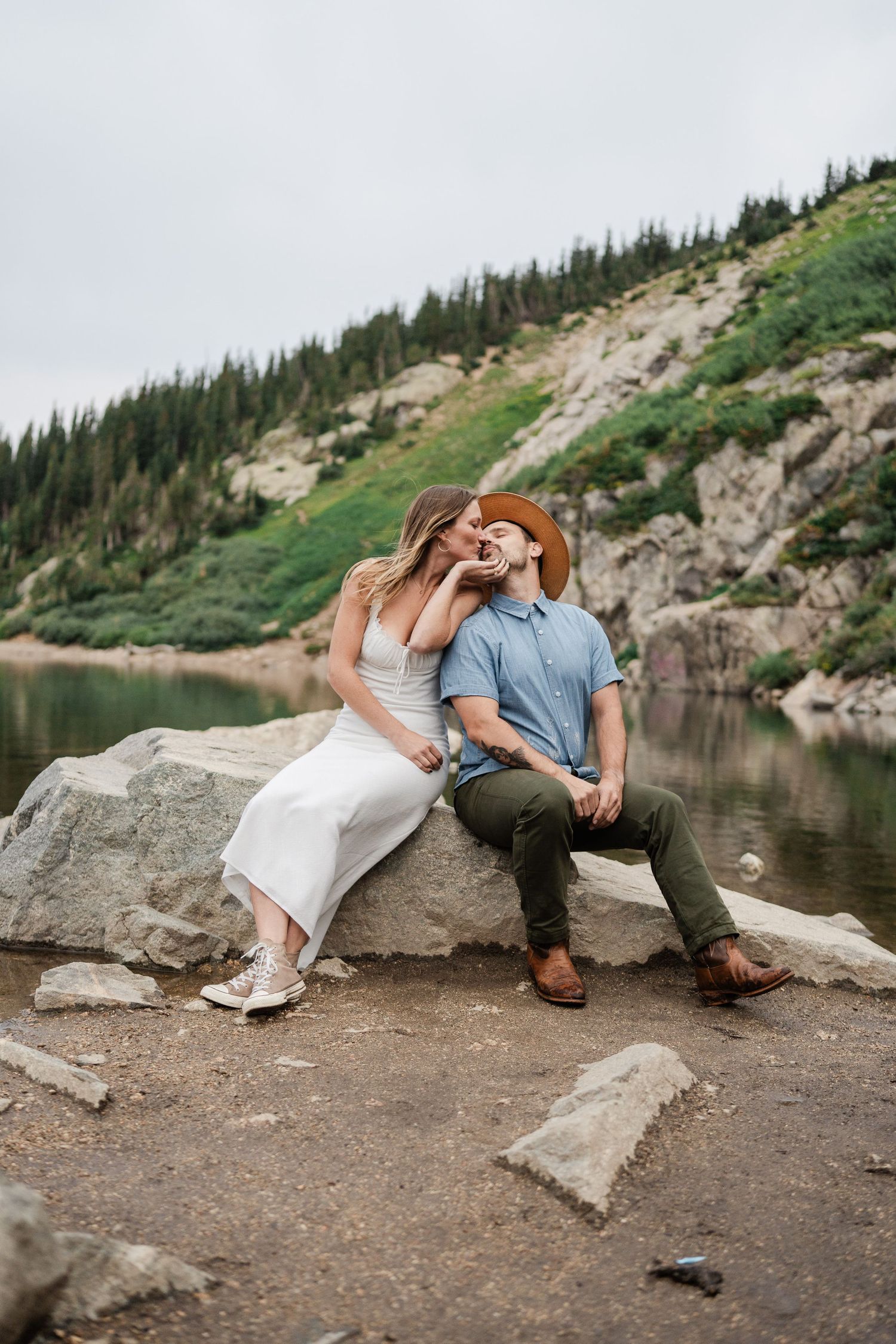 A couple shares a romantic kiss while sitting on rocks beside a serene alpine lake surrounded by pine-covered hills.