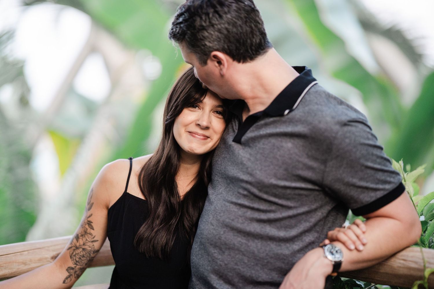 Couple sharing a tender moment outdoors, man kissing woman's head near wooden fence with tropical foliage.