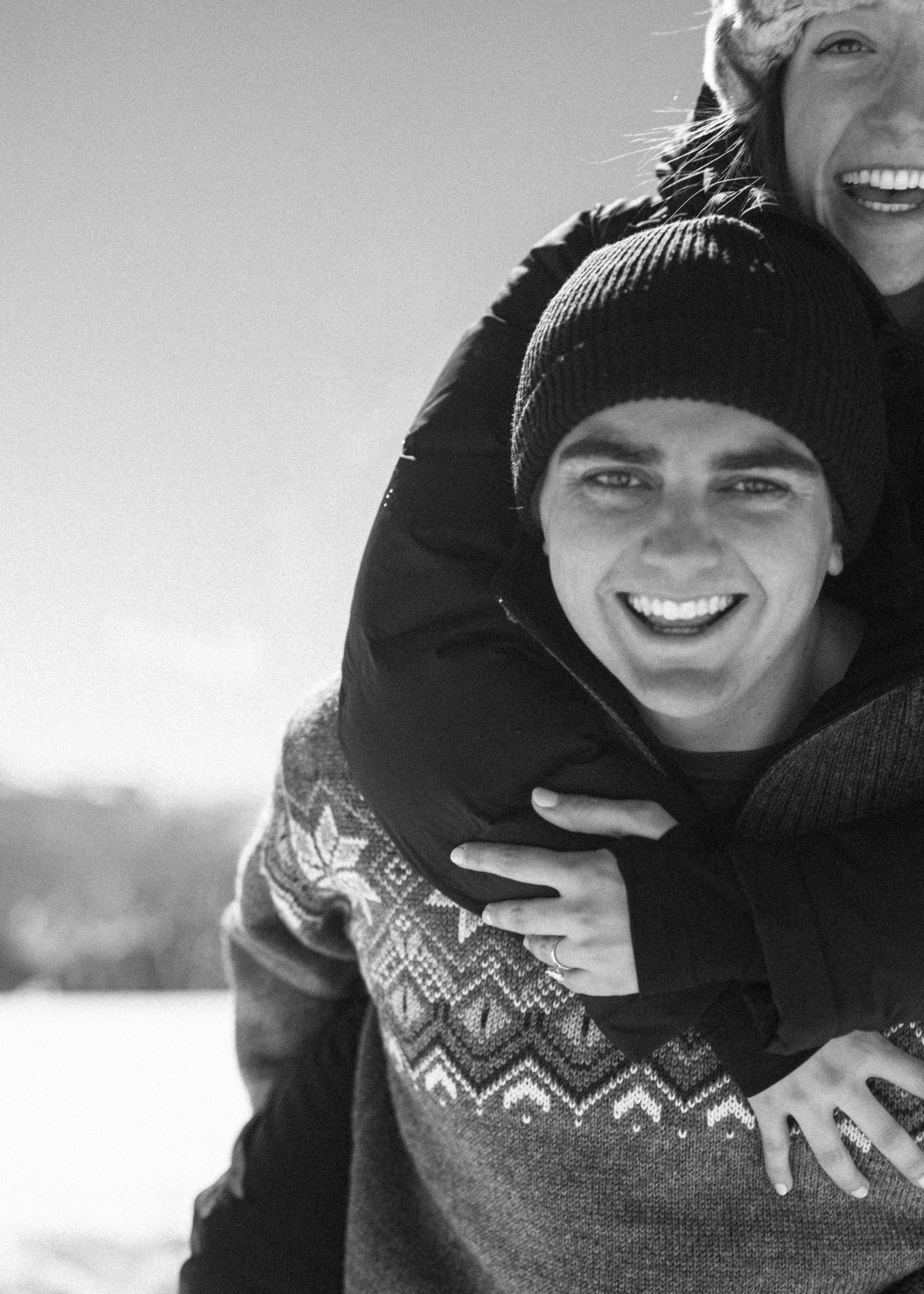 Smiling young man in fair isle sweater gets a piggyback hug from woman in snowy winter setting.
