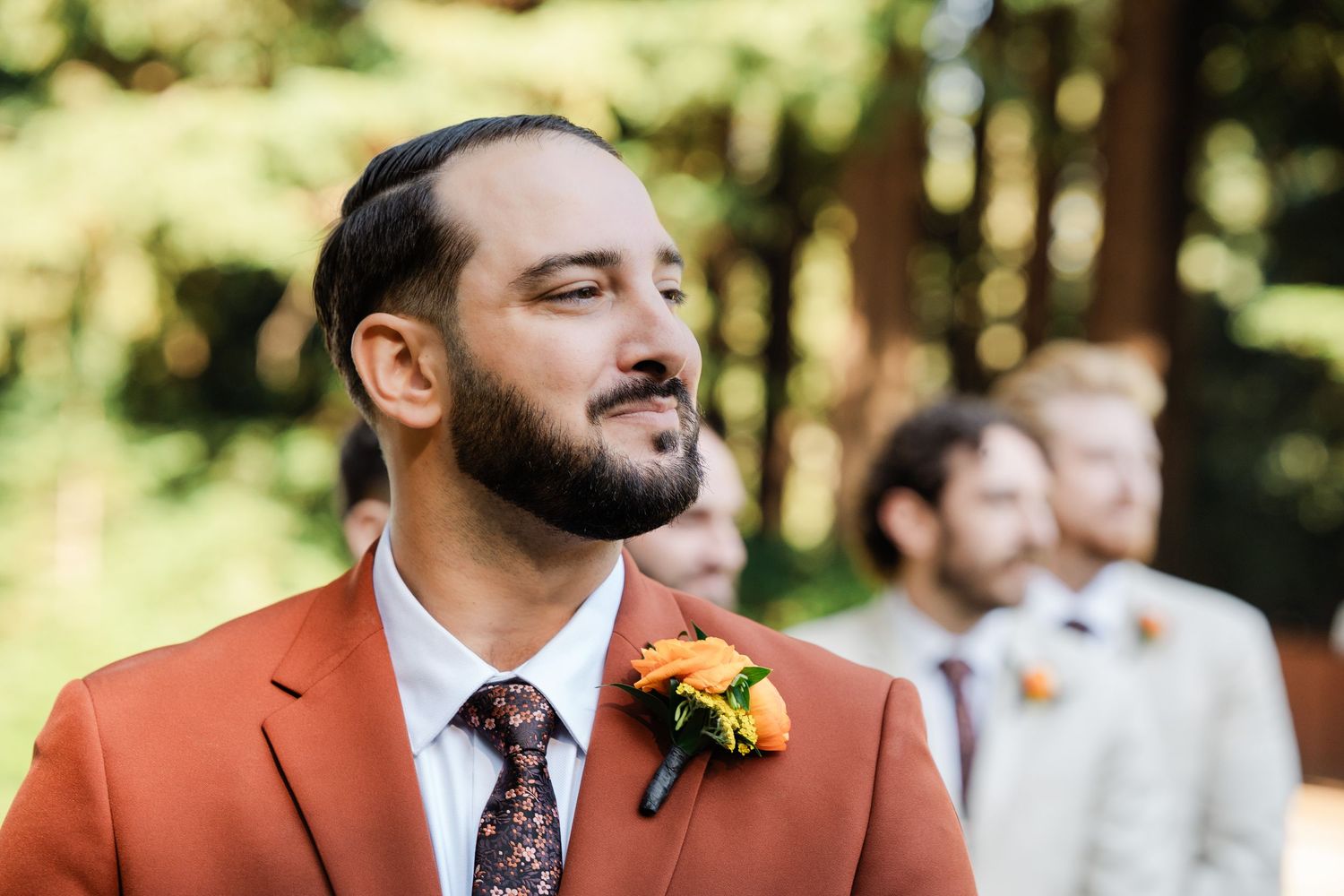 Groom in terracotta suit with orange floral boutonniere gazing during outdoor wedding ceremony.