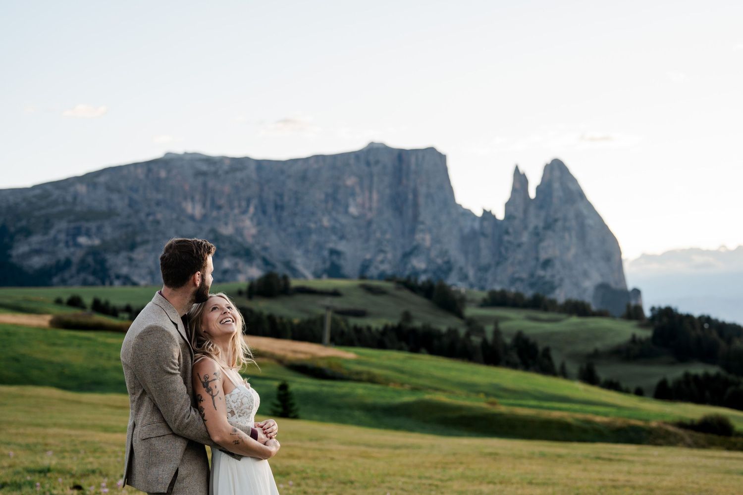 A couple embraces in a lush green meadow with stunning jagged Dolomite mountain peaks rising in the background.
