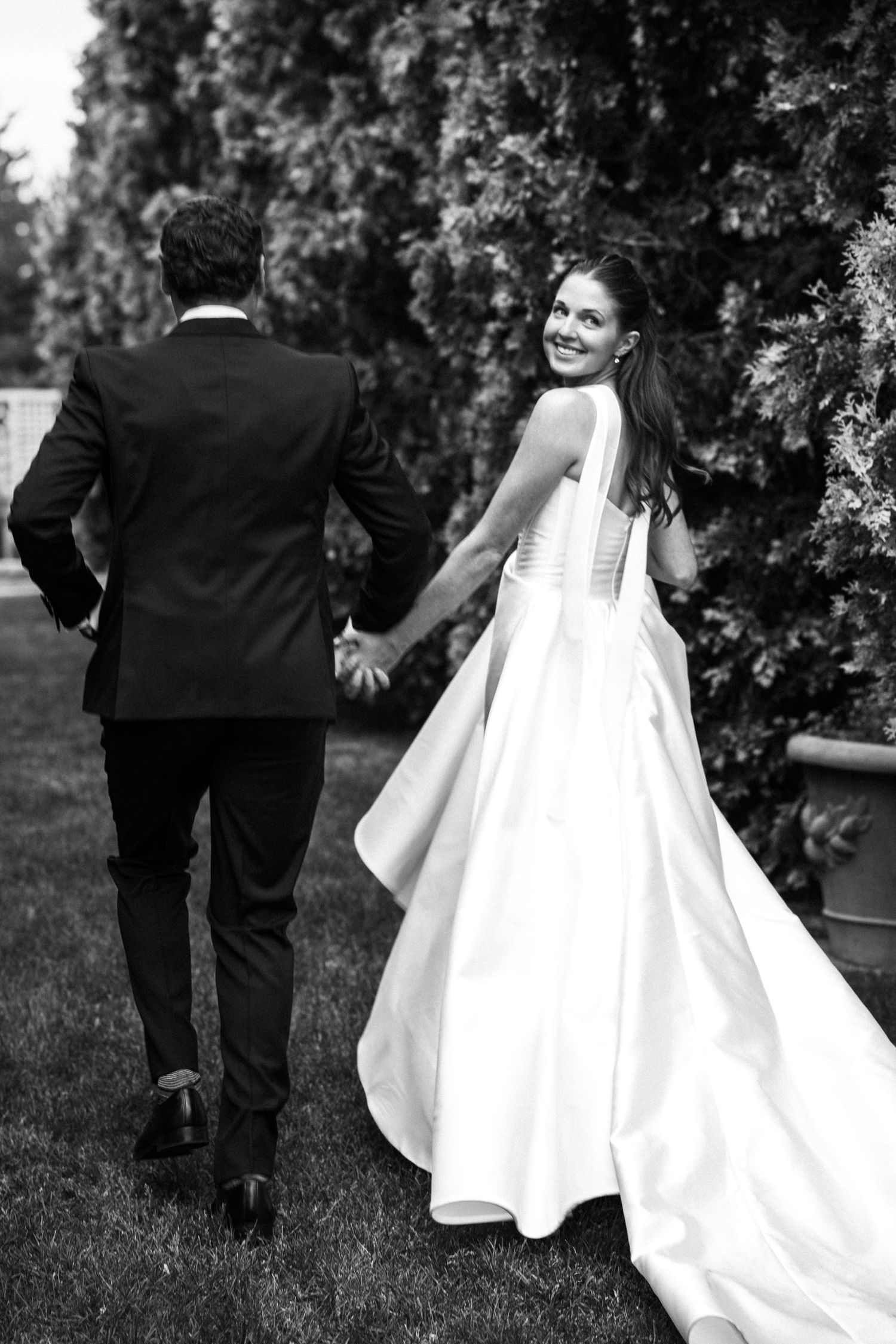 Black and white photo of bride smiling back while holding grooms hand in garden.