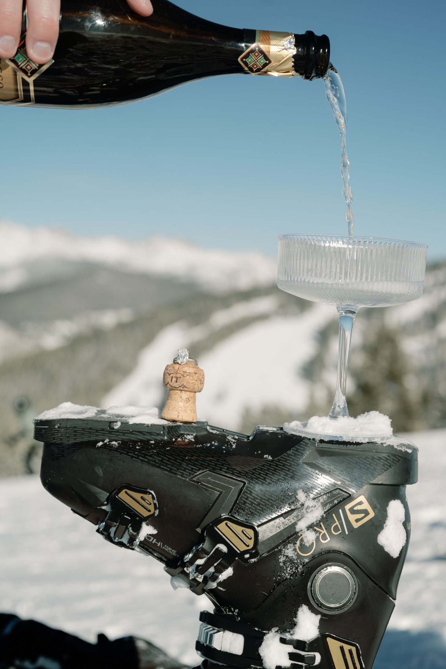 Prosecco being poured into coupe glass balanced on ski boot with snowy mountains behind.