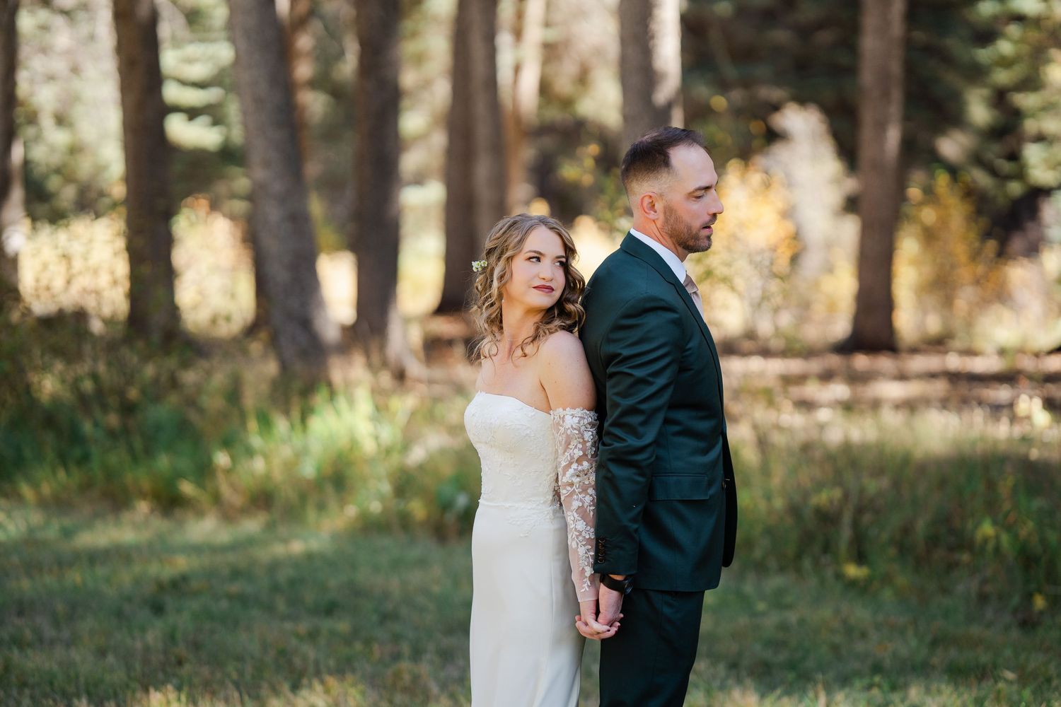 Bride and groom standing back to back in a sunlit forest clearing during wedding portraits.